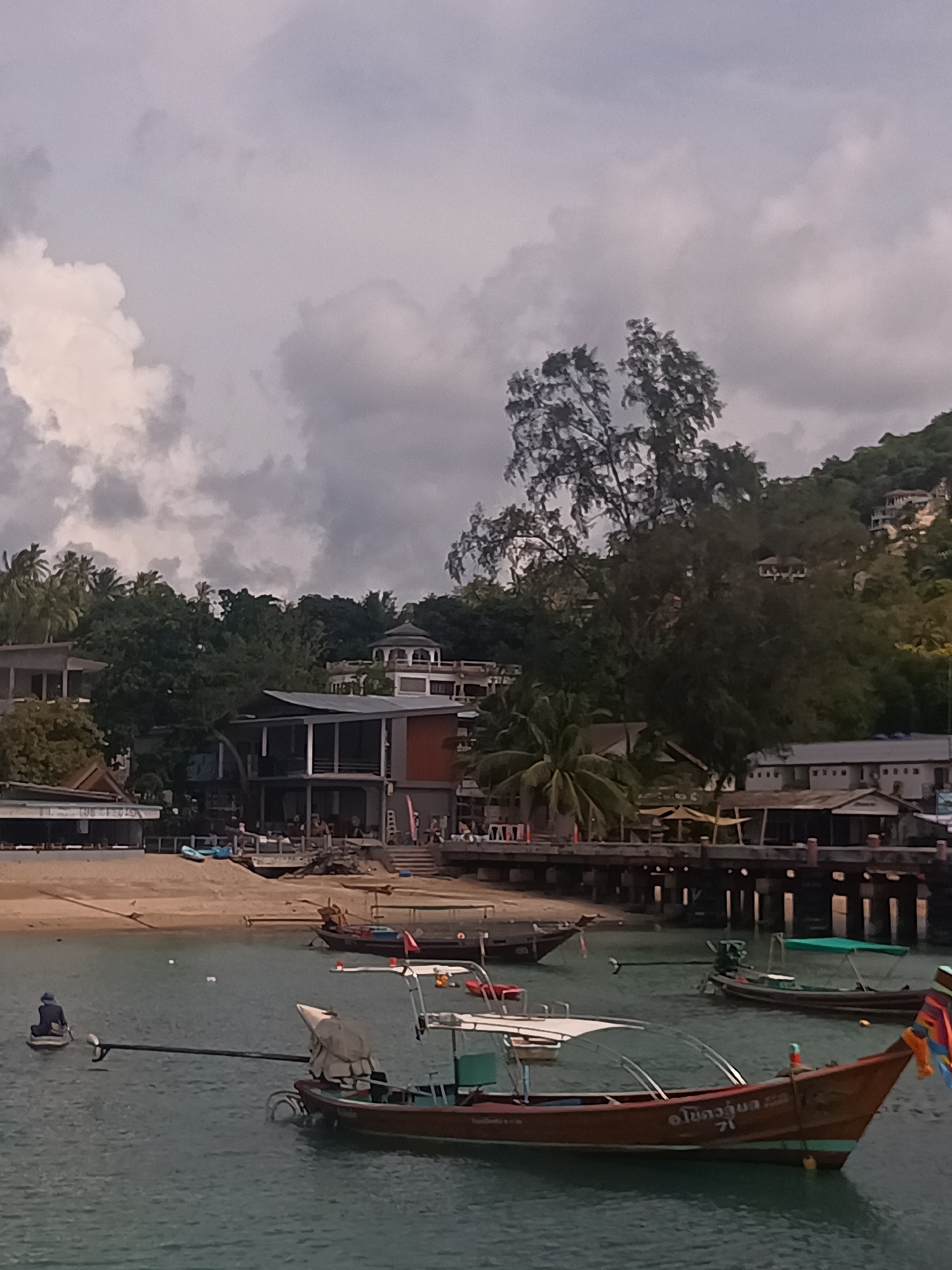 The image depicts a coastal scene in Tambon Ko Tao, Thailand, under a cloudy sky. The foreground features several traditional longtail boats anchored in the water. One boat, predominantly brown in color, has a white canopy, a long tail propeller, and visible Thai script on its hull. In the water, near the left edge of the frame, is a person sitting on a small board. A sandy beach stretches behind the boats, leading to a built-up area with various structures, including multi-story buildings with modern architectural elements. A pier extends into the water, with people visible on the beach and around the pier. The backdrop includes lush green vegetation covering a hillside, with some structures visible among the trees. The overall impression is of a bustling coastal area with a blend of local maritime culture and tourist infrastructure.