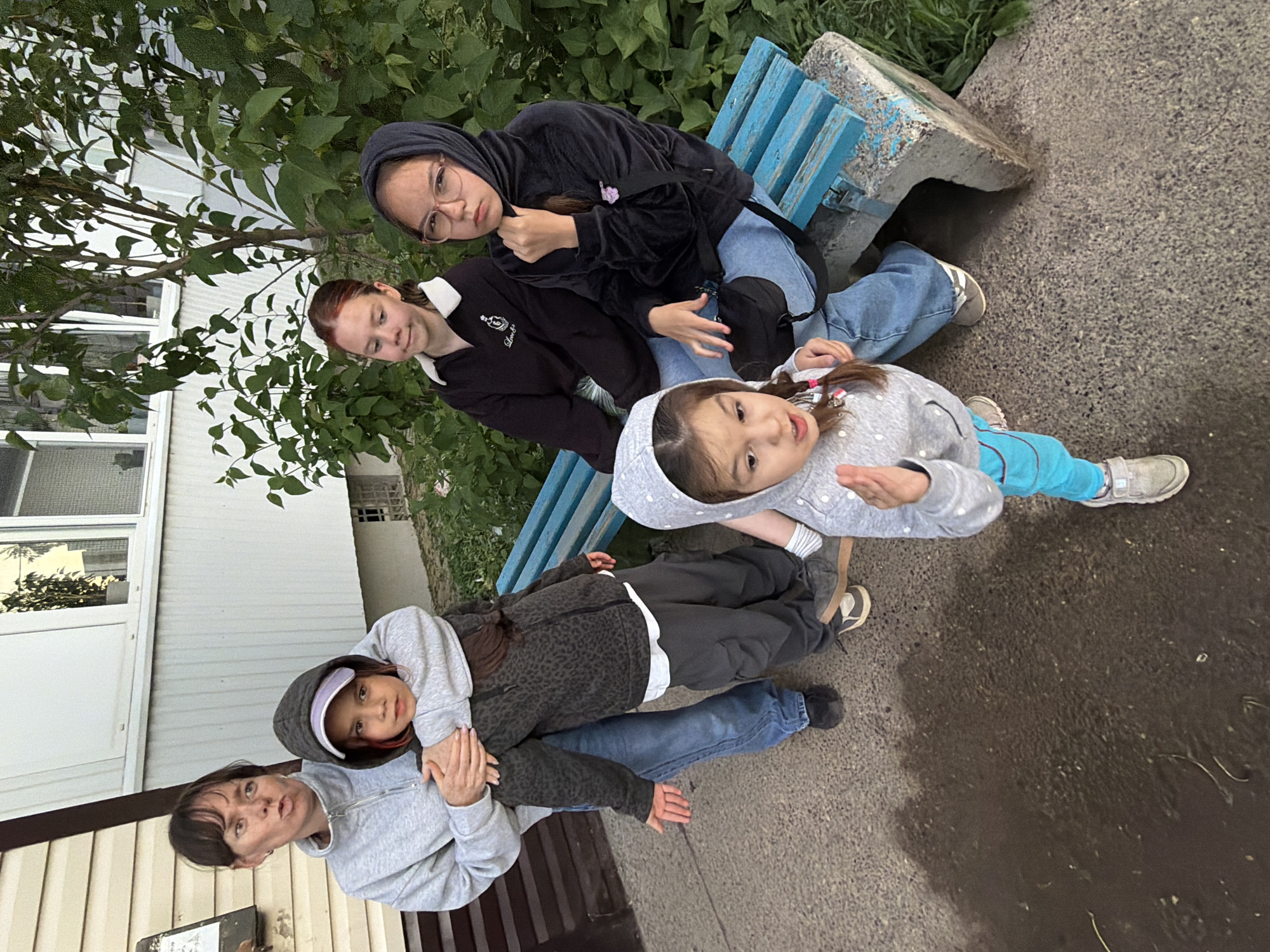This image captures a casual group portrait of five females of varying ages, from young children to adults, posing outdoors in what appears to be a residential or park-like area in Chaikovsky, Russia. The photo is taken from a slightly elevated perspective, with the subjects arranged at different depths.
In the immediate foreground, an adult woman in a light grey zip-up hoodie is making a playful, slightly pouty expression, holding a young child wearing a dark grey patterned hoodie and a light-colored visor. Behind them, partially visible, is another individual (likely an older teenager or young adult) dressed in a black patterned hoodie and blue jeans, appearing to sit or stand lower than the adult.
Further back, seated on a weathered blue wooden bench, is a teenage girl wearing a black hoodie and glasses, looking directly at the camera with a neutral gaze. Next to her, another teenage girl with reddish-brown hair, sporting a black top with a white collar, looks slightly downwards. In the forefront of the image, a younger child in a grey polka-dotted hoodie and bright blue pants is positioned low to the ground, looking up at the camera with wide, curious eyes.
The background features abundant green foliage from trees and bushes, and a section of a light-colored building with windows is visible on the left. The ground is paved with dark, textured concrete or asphalt, showing some dampness. The scene suggests a relaxed gathering during the daytime, likely under an overcast sky due to the soft, diffused lighting. No visible text is present in the image.
