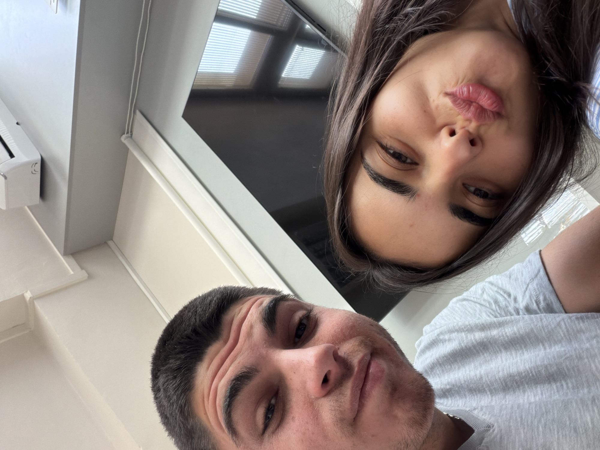 The image is an upward-angled selfie featuring two individuals, a male and a female, set against an indoor background. The male, positioned in the lower-left foreground, has short dark hair, dark eyebrows, and light stubble. He looks upwards with a slightly smirking or relaxed expression while wearing a light grey t-shirt. Above and to his right, the female's inverted face is visible, characterized by long dark hair and dark eyebrows. She is making a playful "pout" face with pursed lips. She wears a light-colored, possibly striped, top.
The background reveals a room with light-colored walls and ceiling. A large, dark flat-screen TV screen is prominent behind the female, reflecting horizontal blinds, which are also directly visible in parts of the background. On the far left, a portion of a white, wall-mounted air conditioning unit is seen, bearing a small, indistinct logo. The ambient lighting suggests the photo was taken during the daytime. The activity is a casual selfie, capturing a moment of playful interaction. While the image itself contains no explicit location markers, it is situated in Sofia, Bulgaria, as per provided context. No discernible text beyond a small, unreadable logo on the AC unit is present.