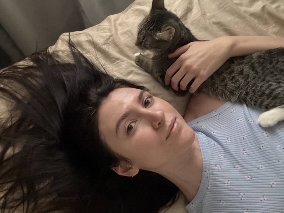 A woman with long, dark hair lies on a tan bedspread, gazing at the camera with a neutral expression. She wears a light blue floral top. A gray tabby cat lies on her, facing away from the camera, with a white paw visible. The woman's hand rests gently on the cat's back. The composition is a close-up, capturing the intimacy between the woman and her feline companion.