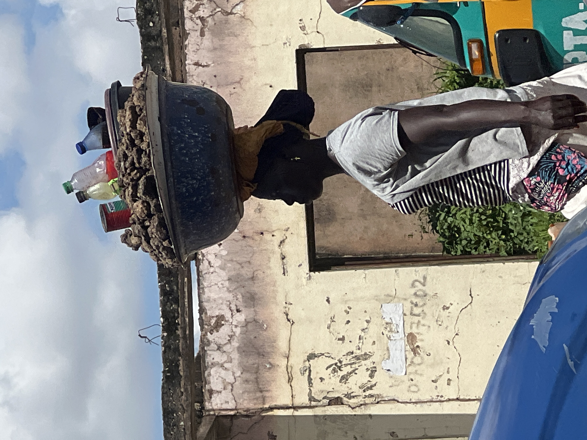 A medium shot shows a woman from the side, walking, with a large blue basin of goods balanced on her head, in Jos, Nigeria. The woman, who has dark skin, is wearing a grey t-shirt over a black and white striped top and a colorful patterned belt. Her hair is pulled back and covered by a yellow cloth. The basin is filled with what appears to be soil or small rocks, with several plastic bottles and a red tin can placed on top. Behind her, a dilapidated building with peeling paint and cracked walls serves as the backdrop. A green and yellow auto-rickshaw is partially visible on the right side of the frame. The sky is cloudy and bright.