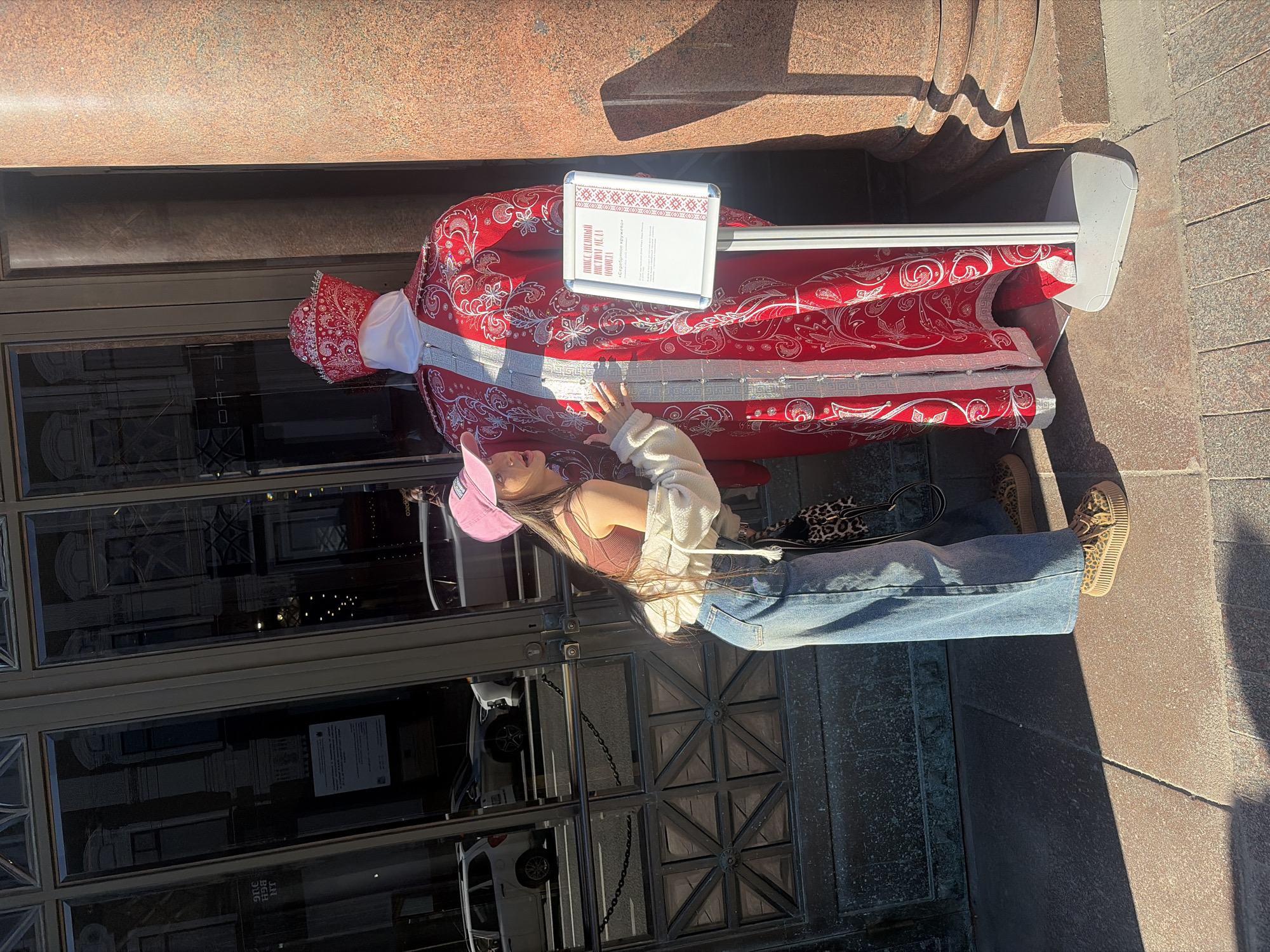 In Saint Petersburg, Russia, a young woman with a pink baseball cap and loose-fitting jeans poses next to a mannequin dressed in a traditional red and white embroidered garment. She is leaning towards the mannequin, with her right hand resting on its sleeve. The mannequin is standing on a sidewalk in front of a building with large glass windows and a granite column. A sign with Russian text and embroidered patterns is attached to a stand beside the mannequin. Cars and reflections of buildings are visible in the glass windows, suggesting an urban environment. The scene is illuminated by sunlight, casting shadows on the ground.
