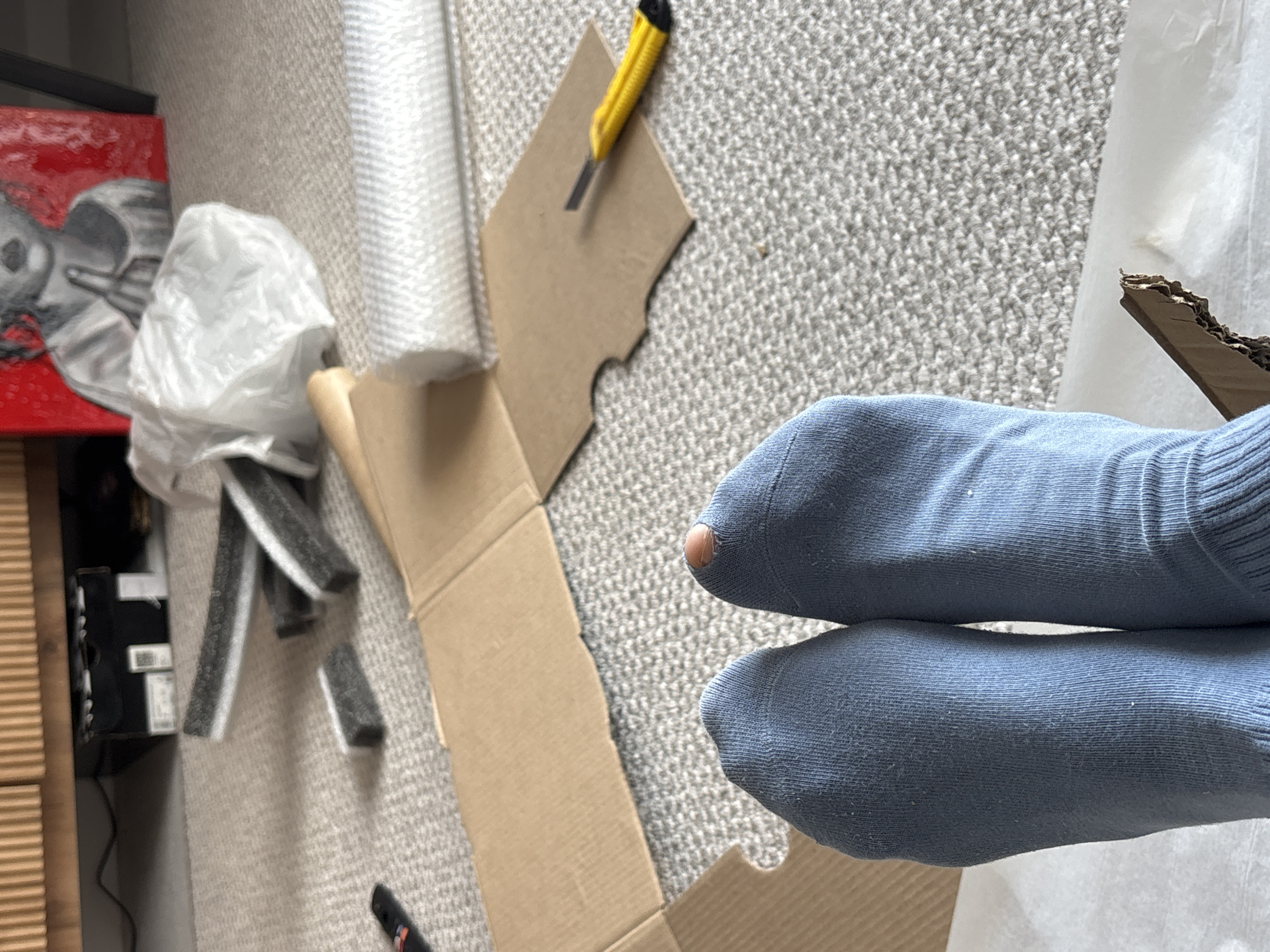 The image shows a close-up, top-down view of a person's feet wearing light blue socks, one of which has a hole revealing a toe. The feet are positioned among packing materials, including cardboard and bubble wrap. A utility knife with a yellow handle lies on a piece of cardboard. In the background, there's a partially visible painting with red and grey tones, along with black foam packaging pieces. The scene appears to be indoors on a carpeted floor. The image contains no clues or evidence connecting the scene to Krasnoyarsk, Russia.