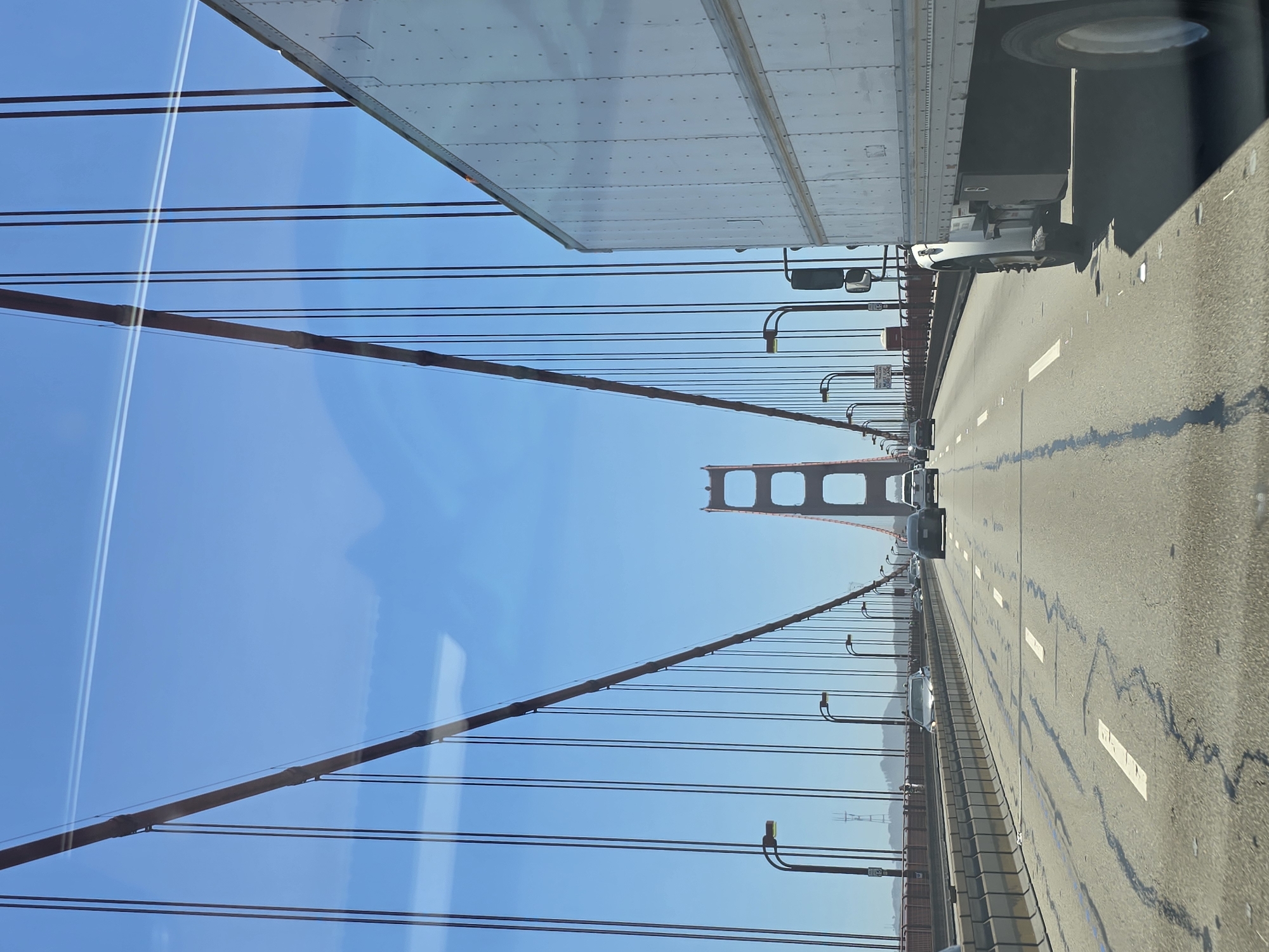The image shows a view from inside a vehicle traveling on a bridge. The bridge is a suspension bridge with large, red towers visible in the distance. The road is paved and has multiple lanes with white lane markings. Several cars and a white box truck are also on the bridge. The sky is blue with some light streaks, possibly from clouds or contrails. The environment appears to be outdoors with a clear day.