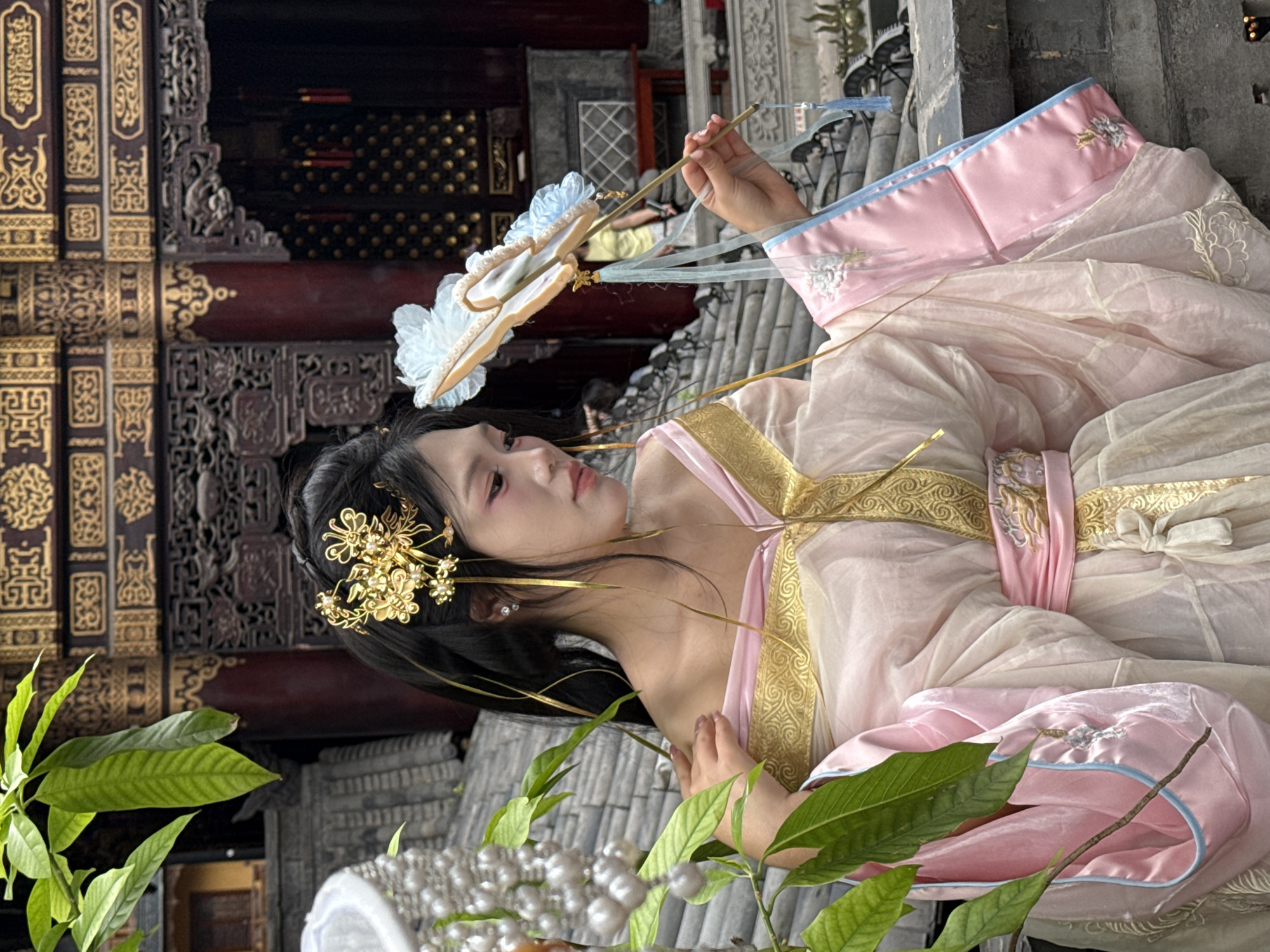 The image captures a woman dressed in traditional Chinese attire in Yuzhong District, China. She is adorned in a light pink hanfu, complete with a golden embroidered belt and hair accessories. She holds a decorative white fan, with blue accents, in her right hand. The setting appears to be an outdoor area near a building with ornate wooden carvings and brickwork, typical of traditional Chinese architecture. There are green plants visible in the foreground. The architecture and attire suggest a cultural or historical context. The grey stone background and the positioning of the subject and plants suggest a deliberate staging for a photoshoot.