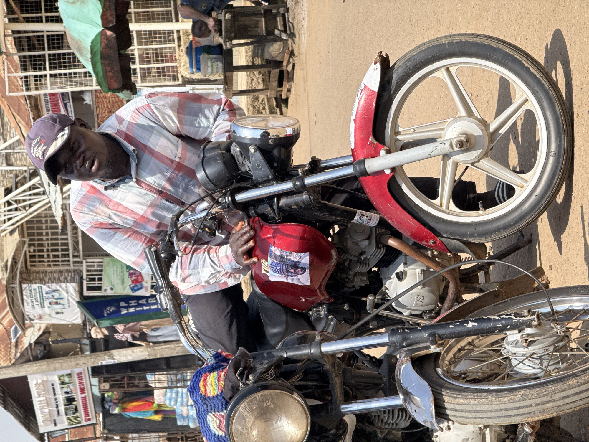 The image shows a man sitting on a red motorcycle in an outdoor marketplace. He is wearing a red and white plaid shirt and a purple baseball cap. The motorcycle has a sticker on its fuel tank featuring a picture of a man. In the background, there are market stalls with various goods, including colorful textiles and what appear to be bags of cement. The scene suggests a bustling commercial area in Abuja, Nigeria.