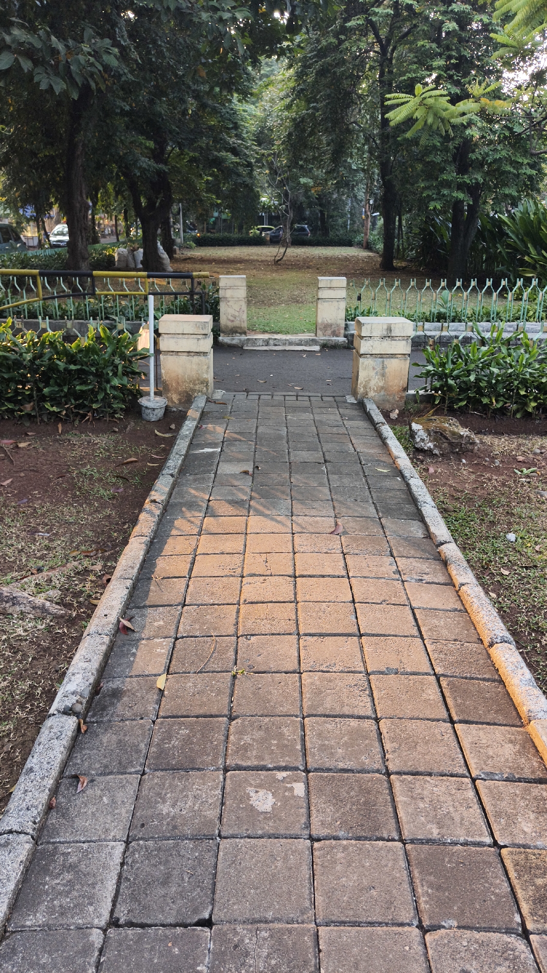 A paved walkway of rectangular grey stones leads through a park in South Jakarta, Indonesia. The walkway is flanked by a low green hedge on the right and a mix of dry grass and soil with some fallen leaves on the left. Two stone pillars stand at either side of the walkway, marking an entrance to a grassy area beyond. Trees with lush green foliage line the background of the park, with some vehicles visible in the distance. The sun casts warm shadows across the walkway, indicating it is likely late afternoon. The overall scene depicts a serene park environment.