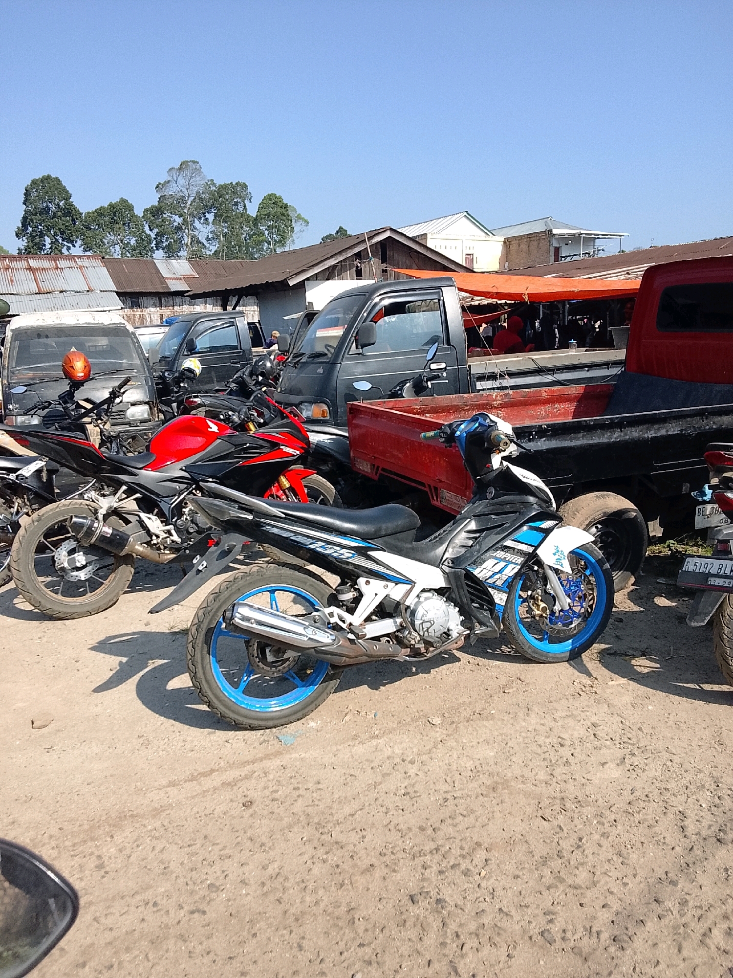 In Bahway, Indonesia, a sunny day is captured with a clear blue sky overhead. The scene is a dirt lot filled with various vehicles, suggesting a marketplace or transport hub. Several motorcycles, including a red Honda and a white and blue sport bike, are parked, some with their wheels turned. Behind them, a row of utility trucks is visible. A black truck with a red flatbed is prominent in the center, and a red truck with a black cargo bed is on the right. The buildings in the background appear to be a mix of corrugated metal and wood structures, with some featuring orange tarps, hinting at temporary stalls or coverings. The overall impression is one of activity and commerce in a somewhat rural or industrial area.
