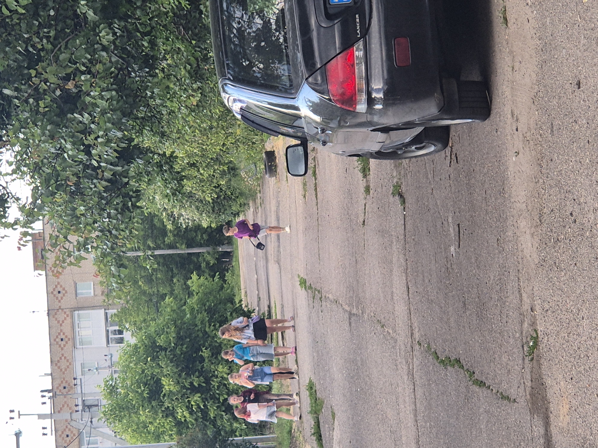 In the city of Sozonivka, Ukraine, a group of five young females is observed walking on a paved road. They are positioned towards the left side of the image, with their backs mostly to the camera. One female, further to the right and slightly ahead of the group, is walking in the same direction. This individual is wearing a purple shirt and grey shorts, and is carrying a black bag. A black car, identified as a Lancer by the visible badging, is parked on the right side of the road, partially obscuring the view of the right edge of the scene. The environment is characterized by dense green trees lining the right side of the road, with a building visible in the background on the left. The road surface appears weathered and cracked, with grass growing in some of the crevices. The sky is overcast.
