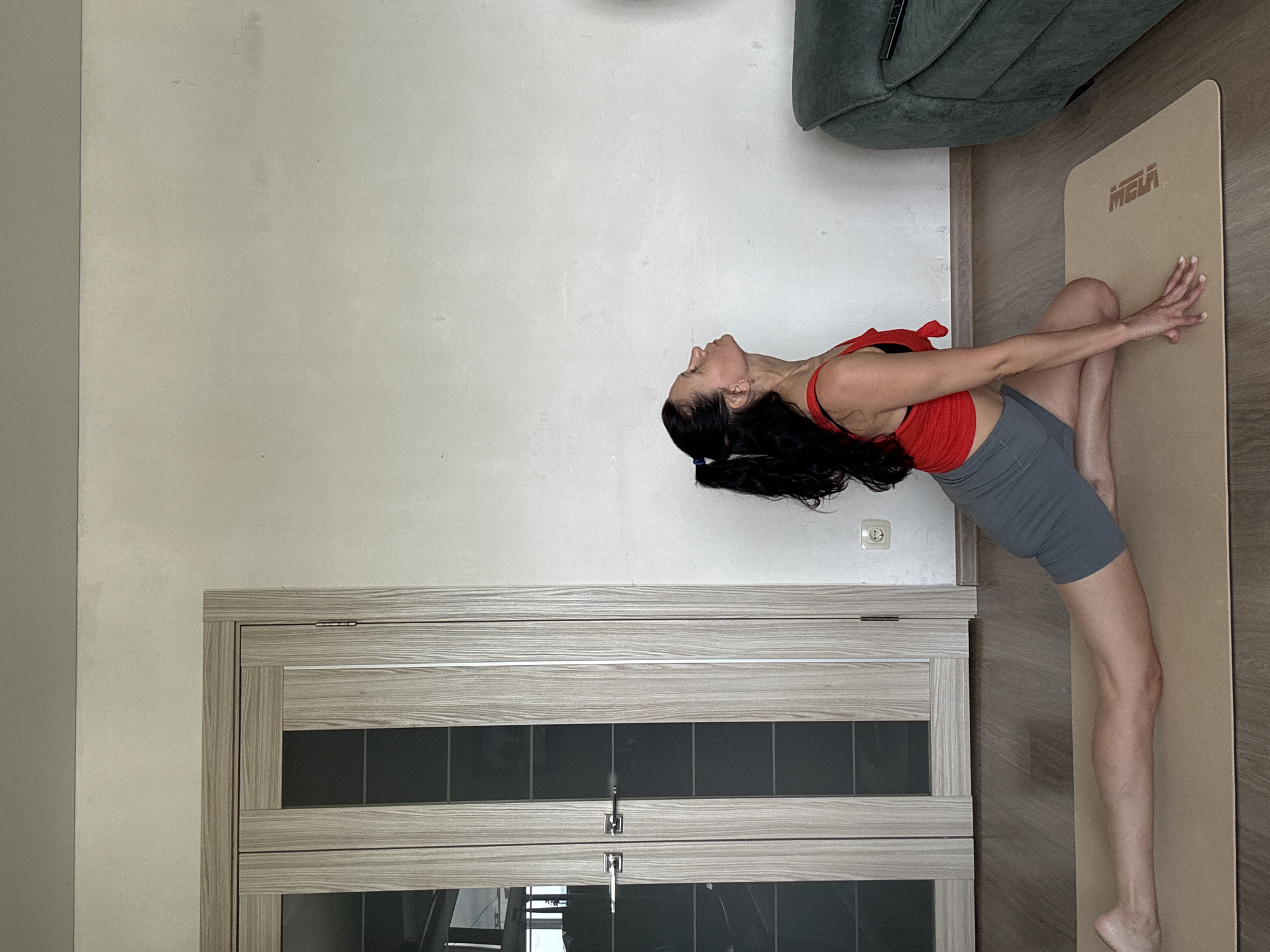 A woman with dark hair pulled back in a ponytail, wearing a red tank top and grey shorts, is performing a yoga pose on a tan yoga mat. She is positioned on a wooden floor in front of a large set of double doors with frosted glass panels and silver handles. The doors are light brown and appear to be made of wood with a grain pattern. The walls are plain and white. To the right of the woman, a portion of a green sofa is visible. The scene appears to be indoors, likely in a residential setting, but there is no specific indication of the city of Chaikovsky, Russia.