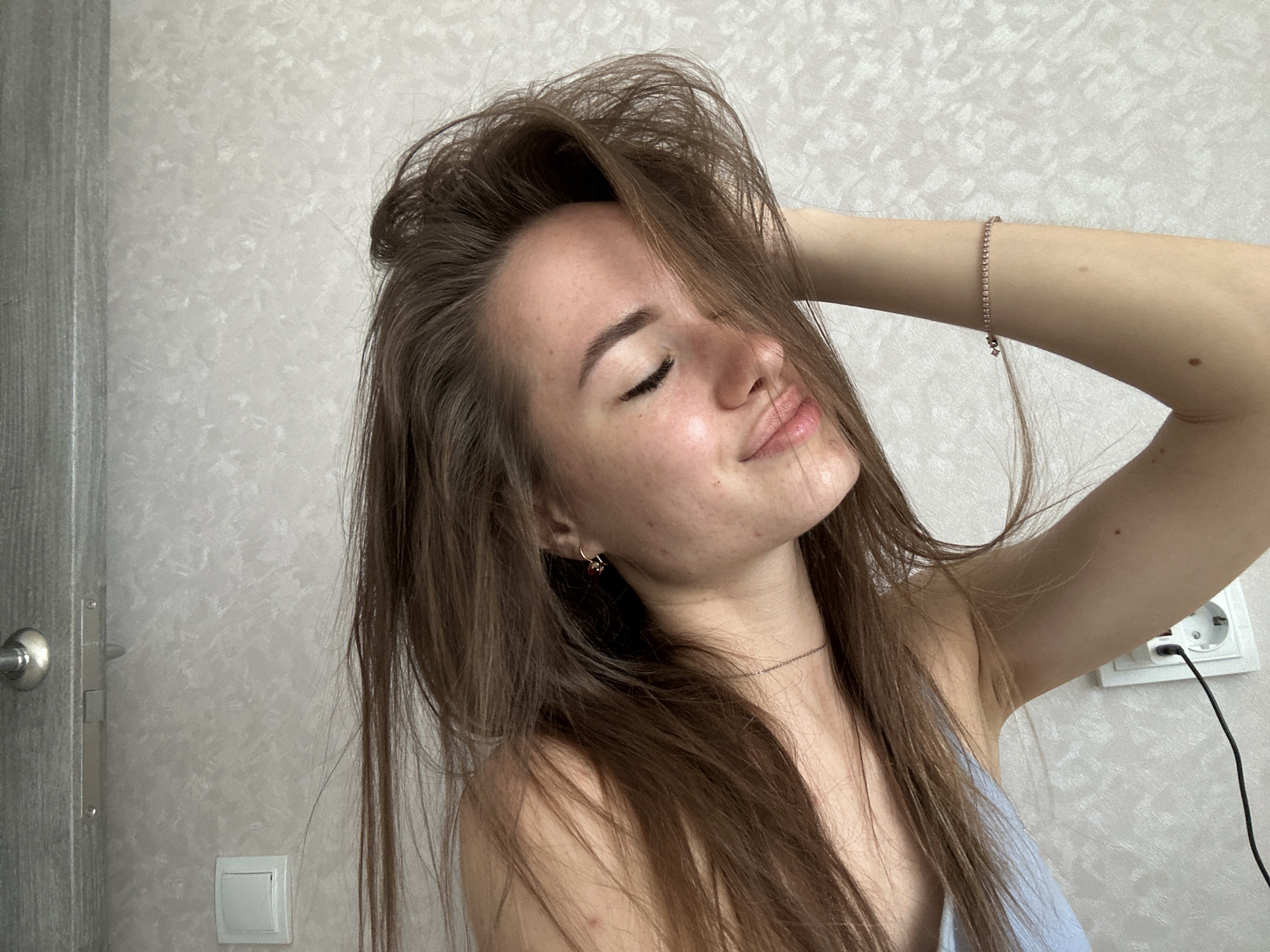 A medium close-up shot captures a young woman indoors, posing with her eyes closed and a slight smile or pout on her lips. She has long, messy brown hair that frames her face and is lifted by her right hand, which is raised and possibly resting on her head. A delicate, sparkly bracelet adorns her right wrist. She wears a thin chain necklace and a small hoop earring in her visible left ear. Her light-colored top appears to be a camisole or tank top.
The background features a light grey or off-white textured wallpaper. To the left, part of a light-colored wooden door with a silver handle is visible. To the right, a white power outlet with a black cable plugged in, and a white light switch are mounted on the wall. The lighting is bright and natural, suggesting it is daytime. The woman's face shows some freckles and skin imperfections, adding to a natural appearance. Her expression conveys a sense of calm or contemplation. No visible text is present in the image.