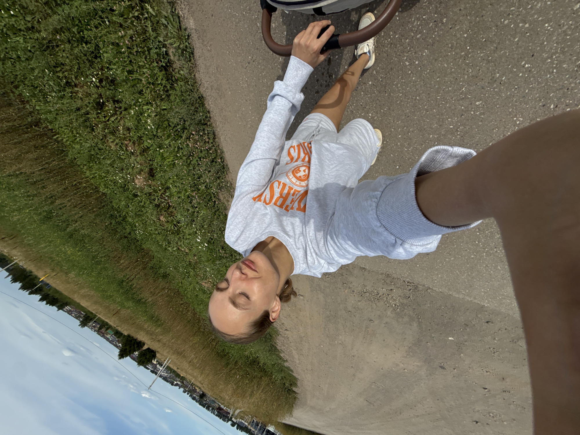 The image shows a woman from a first-person perspective, holding a stroller. She is wearing a light grey sweatshirt with orange lettering and grey shorts. Her eyes are closed, and her face is tilted towards the sun, suggesting she is enjoying the warmth. She is standing on a paved path that appears to be in a rural or suburban area. To her left, there is a field of dry grass and some sparse vegetation. In the background, there are trees and houses, indicating a settlement. The sky is blue with some scattered clouds. The stroller is partially visible on the right side of the frame, with a brown handle. The overall scene appears to be a daytime outdoor activity in a peaceful setting, possibly in or near the village of Podshivalovo, Russia, given the visual cues of rural landscape and residential buildings.