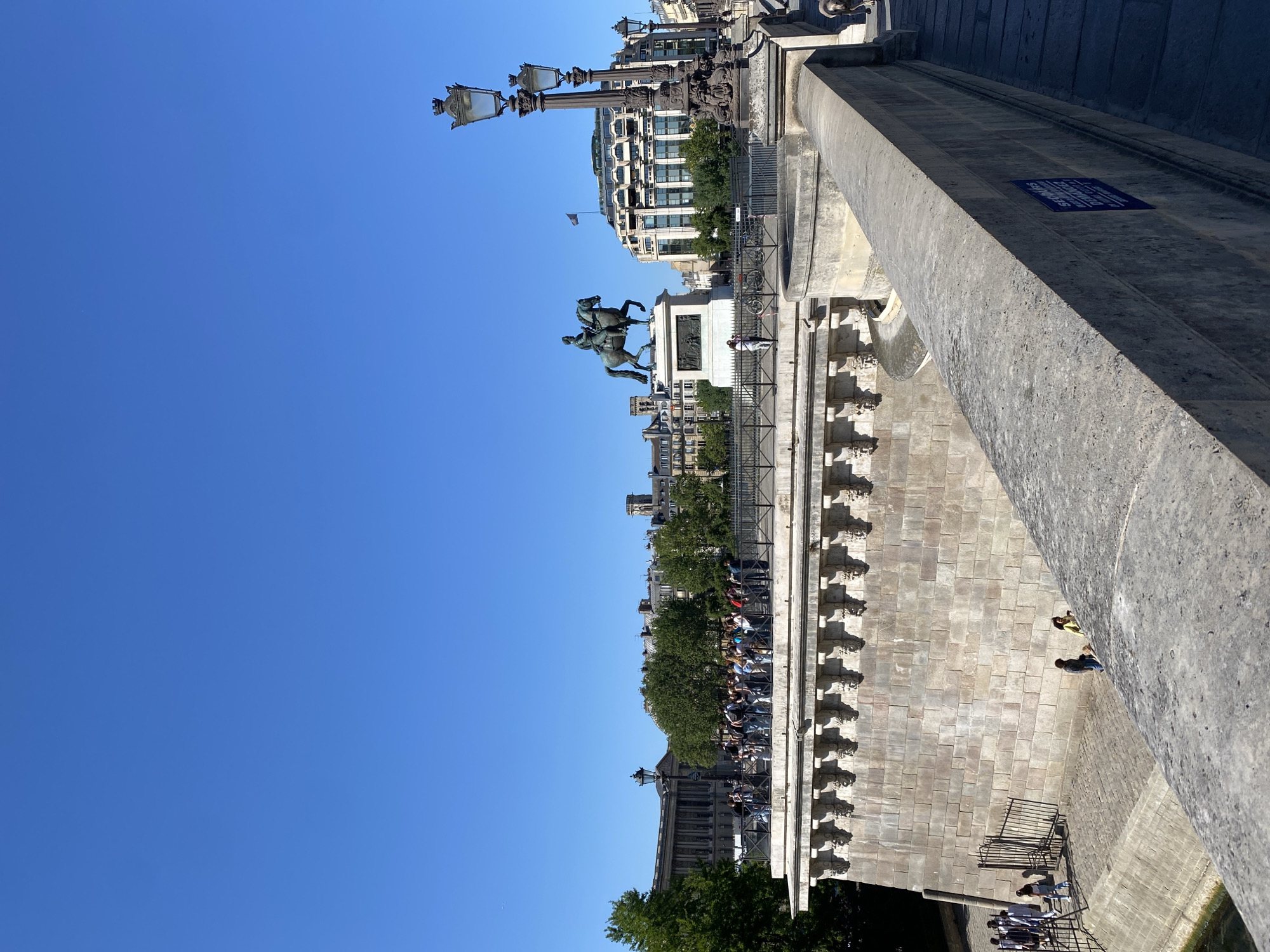 The image, taken on a sunny day in Paris, France, captures a view across a historic stone bridge or embankment. The sky is a vibrant, clear blue, indicative of fair weather.
In the mid-ground and right, a substantial grey stone balustrade or wall of the bridge stretches diagonally, featuring a small blue sign affixed to its surface. The sign displays French text: "SE DEMANDER SI LES PARISIENS SONT TRISTES QUAND IL PLEUT" (To wonder if Parisians are sad when it rains).
Centrally located, a prominent bronze equestrian statue stands atop a stone pedestal. The statue depicts a man on horseback, with ornate, multi-lamp streetlights flanking the area. Behind the statue, a metal railing defines the edge of the bridge's upper walkway, separating it from the views below. A crowd of people is gathered along this railing, appearing to observe the surroundings or simply enjoying the day.
In the background, beyond the bridge, classic Parisian buildings with multiple stories and traditional architecture are visible amidst green trees, all bathed in bright sunlight. Below the main bridge structure, on a lower embankment or walkway, several other individuals are present, some near steps leading to different levels. The bridge itself incorporates decorative stone carvings along its lower facade, adding to its historical character. The overall scene conveys a lively yet relaxed atmosphere of urban sightseeing and leisure on a beautiful day.