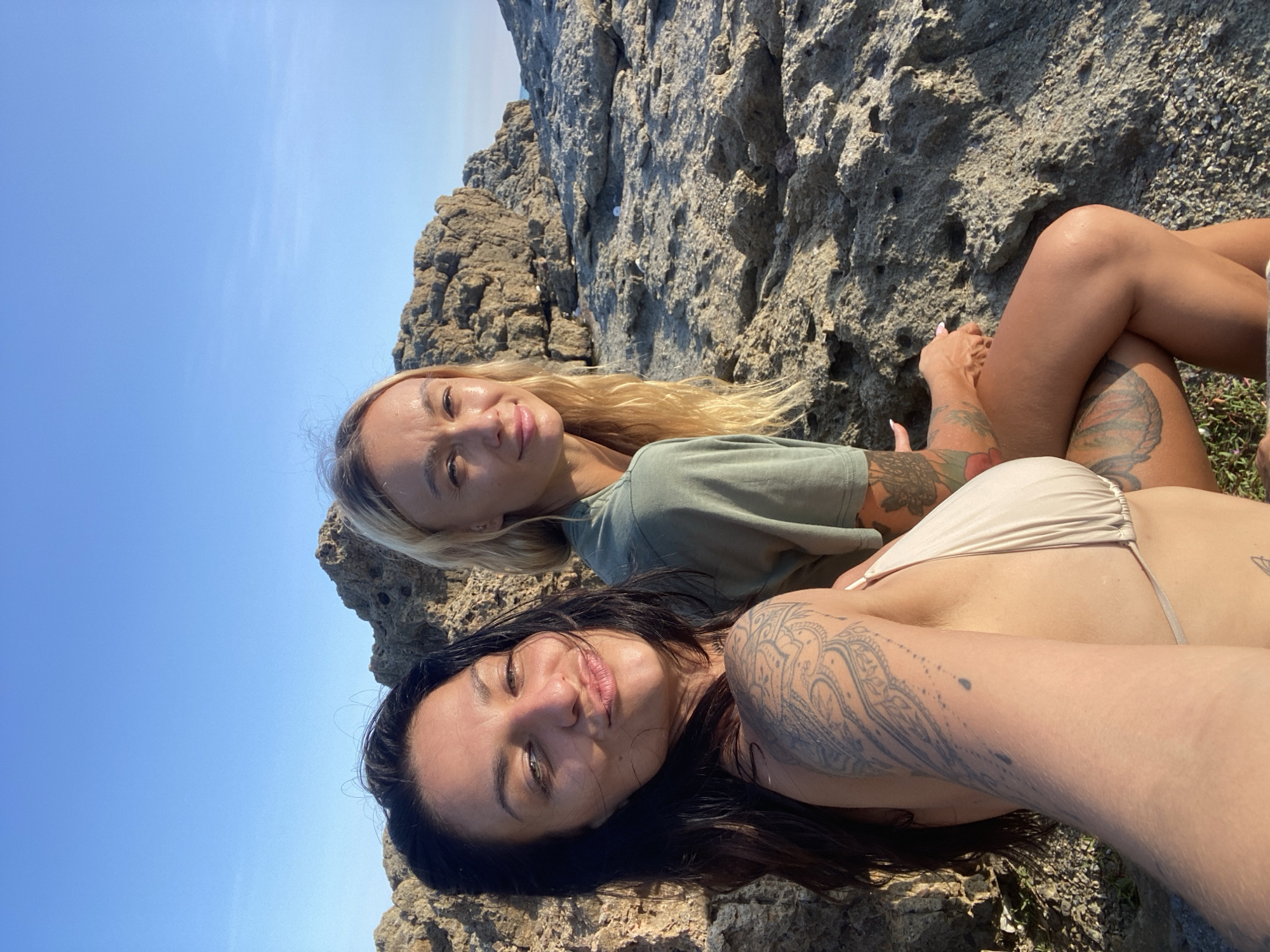 Two women are posing for a selfie on a rocky beach under a clear blue sky. The woman in the foreground on the left has dark hair and is wearing a light-colored bikini top. She has extensive tattoos on her left arm and shoulder. She is puckering her lips. The woman on the right has blonde hair and is wearing a green t-shirt. She also has tattoos on her arms and legs. They are both sitting on rocks that appear to be part of a cliff face. The background shows more rocks and the ocean in the distance. The lighting suggests it is daytime, possibly late afternoon due to the warm tones.