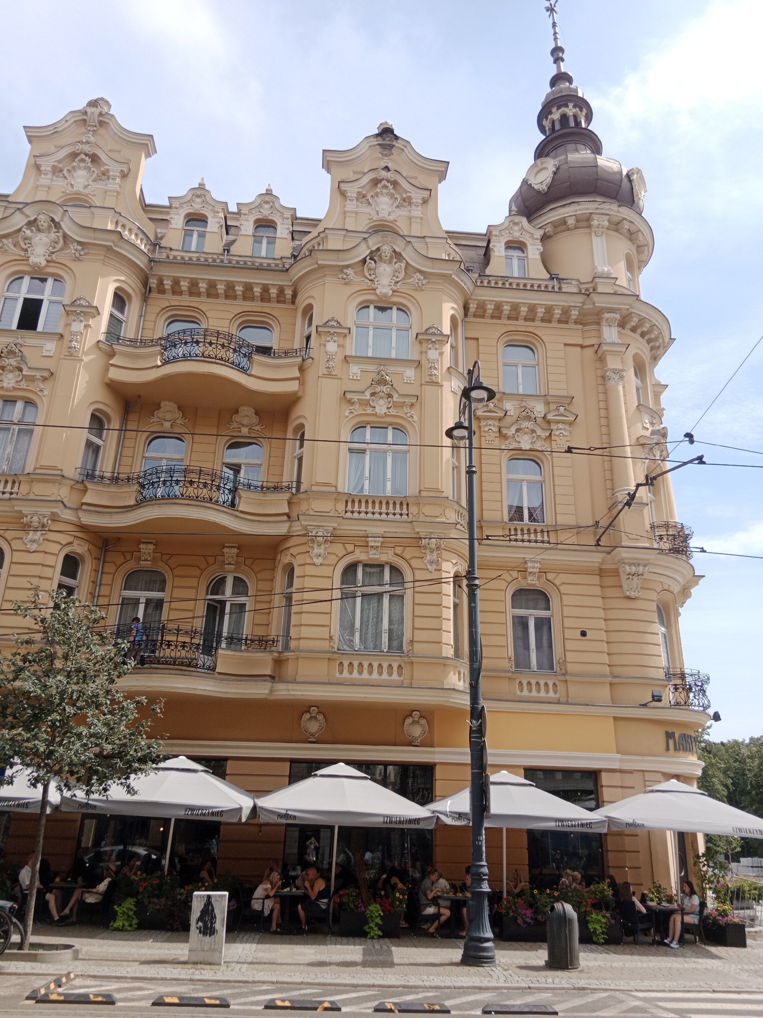 The image depicts the facade of a grand, ornate yellow building in Bydgoszcz, Poland. The building features multiple stories with elaborate architectural details, including curved balconies with wrought-iron railings, decorative carvings around windows and cornices, and a prominent domed tower. There are several windows visible on the facade, some with white curtains.
In the foreground, a tree is visible on the left, and a street lamp stands centrally. Outside the building, at street level, is a covered outdoor seating area for a cafe or restaurant. Several large grey umbrellas provide shade over tables where patrons are seated, engaged in conversation and eating. People of various ages are seated at different tables. Flowers are planted in planters around the seating area. The street in front of the cafe has crosswalk markings. Electrical wires are strung across the upper portion of the image, indicating this is likely a street with tram or trolleybus service. The sky is partly cloudy.
The building's inscription "MARKET" is visible on the ground floor. This suggests the building is located in the Market Square area of Bydgoszcz.