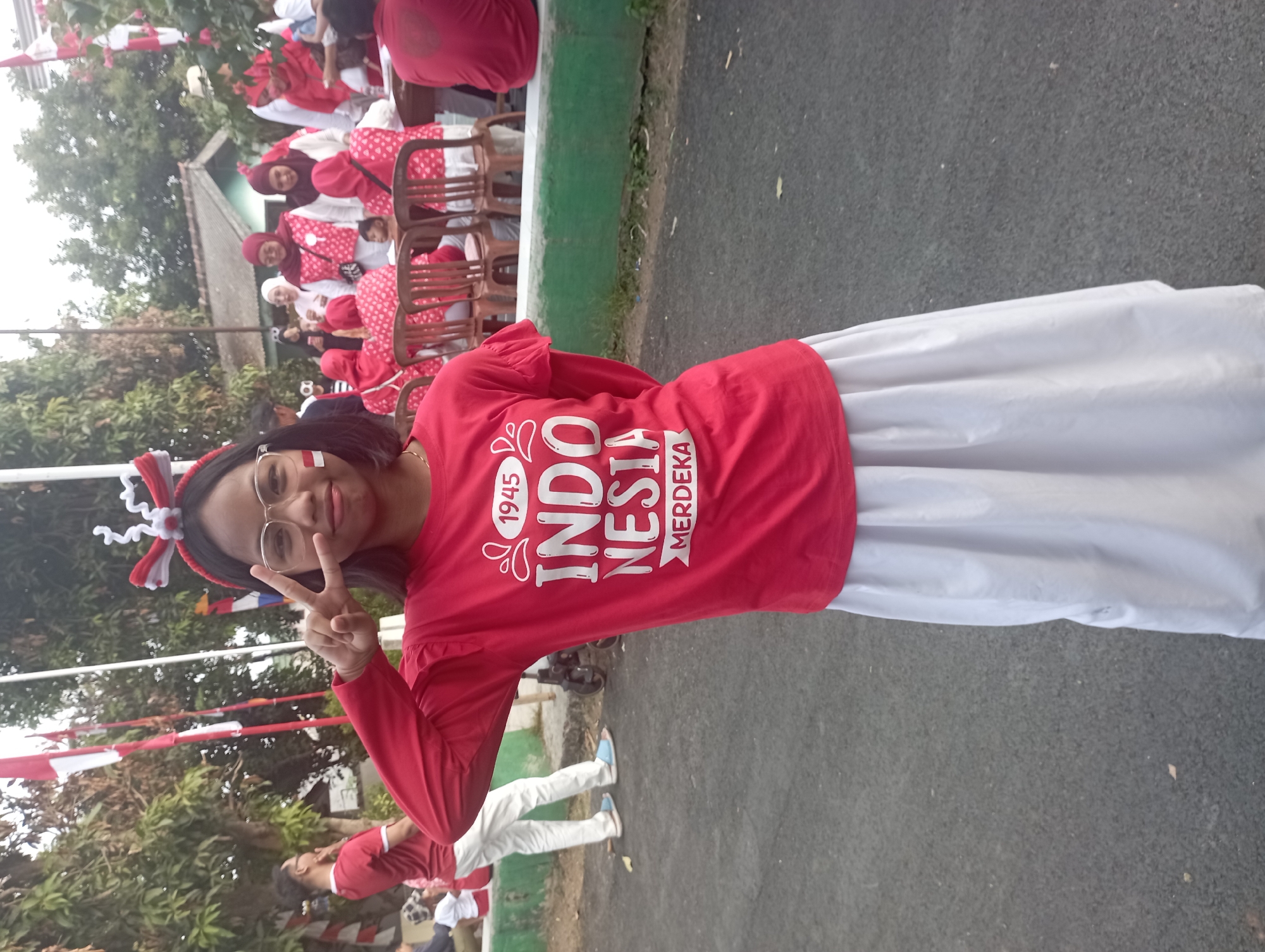 The image shows a young girl in the foreground, standing and posing for the camera. She is wearing a red long-sleeved shirt with "1945 INDONESIA MERDEKA" printed on it, and a white skirt. She has a red and white headband with white fluffy adornments on her head, and a red and white stripe painted on her left cheek. She is wearing glasses and making a peace sign with her right hand. In the background, several people are visible, some wearing red and white attire, suggesting a celebratory or patriotic event. There are also trees, flags (likely the Indonesian flag), and chairs in the background. The setting appears to be outdoors, possibly a street or a public gathering area. The presence of Indonesian flags and the text on the shirt strongly suggest the location is Indonesia, and the clothing and festive atmosphere point towards an Independence Day celebration or similar nationalistic event. Based on the context of the prompt, the location is identified as Purwakarta, Indonesia.