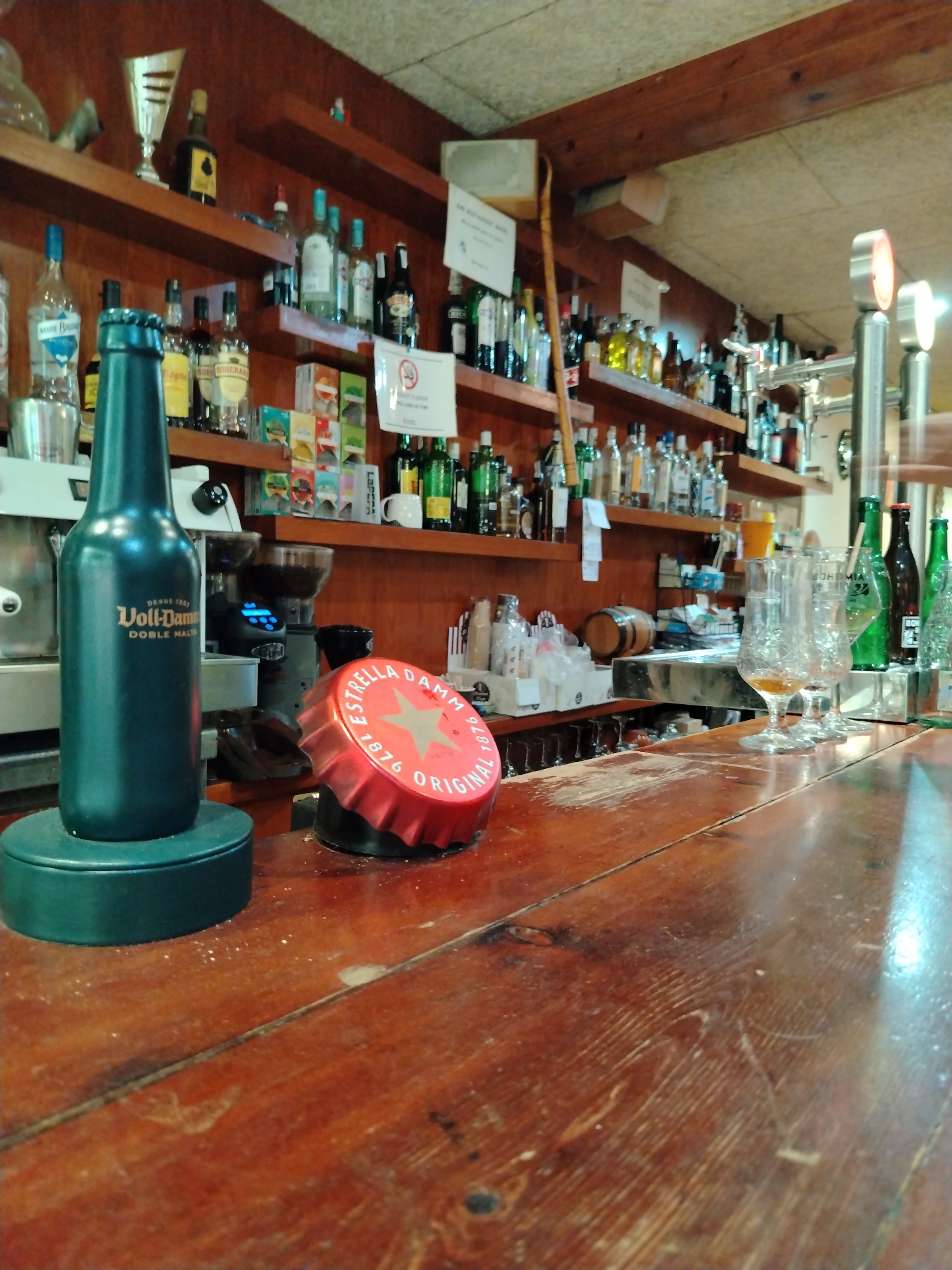 This image shows a close-up view of a wooden bar counter in a bar, likely in Hostalric, Spain. In the foreground, a large, dark green bottle with the label "Voll-Damm DOBLE MALTA" stands on a circular dark green base. Next to it is a red bottle cap that reads "ESTRELLA DAMM 1876 ORIGINAL" with a star logo. The bar counter is made of worn, polished wood with visible grain and some scuff marks. In the background, shelves are stocked with various bottles of liquor, glasses, and bar equipment, including a coffee grinder with a blue digital display. Two empty glasses with some residue are on the bar, and behind them are several bottles of beer and other spirits. The overall impression is of a well-used, possibly traditional, bar setting.