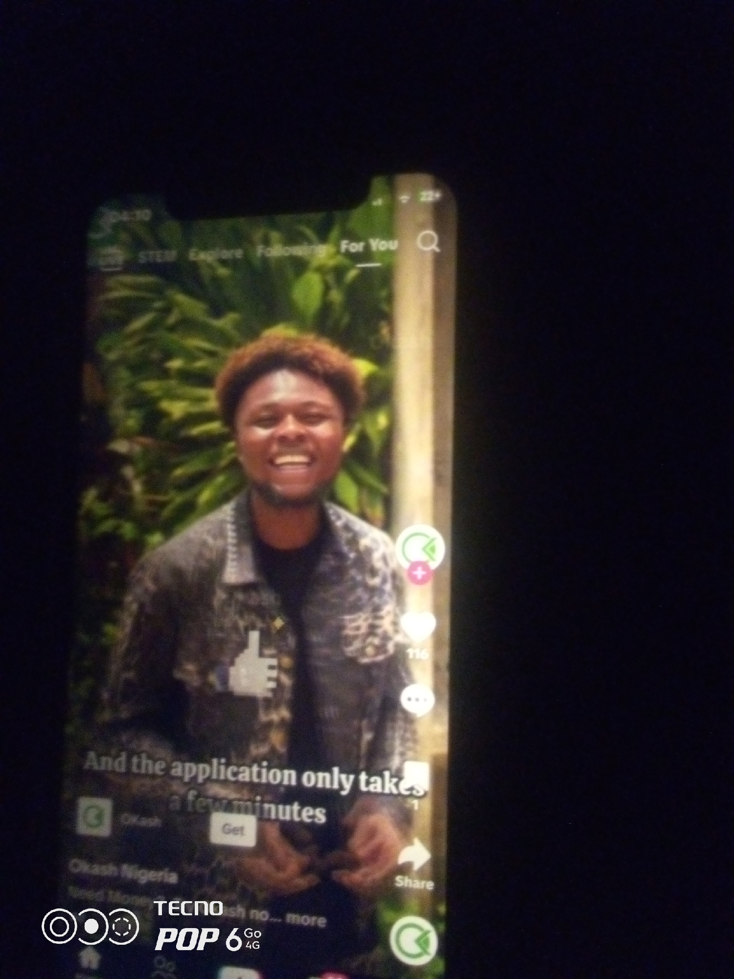 A close-up shot of a smartphone displays a video of a young Black man with curly hair and a beard, smiling broadly. He is wearing a dark jacket with a light-colored pattern. The video appears to be a short-form content platform, indicated by icons like a heart, a speech bubble, and a share arrow on the right side of the screen, along with text at the bottom that reads "And the application only takes a few minutes." Below this, the text "Okash Nigeria" and "TECNO POP 6 4G" are visible, suggesting the phone model and a potential app being featured. The background of the video shows lush green foliage. The smartphone itself is held in a dark environment, likely indoors or at night, with the screen being the primary source of light. The specific location in the city of Howayi, Nigeria, is not determinable from the image.