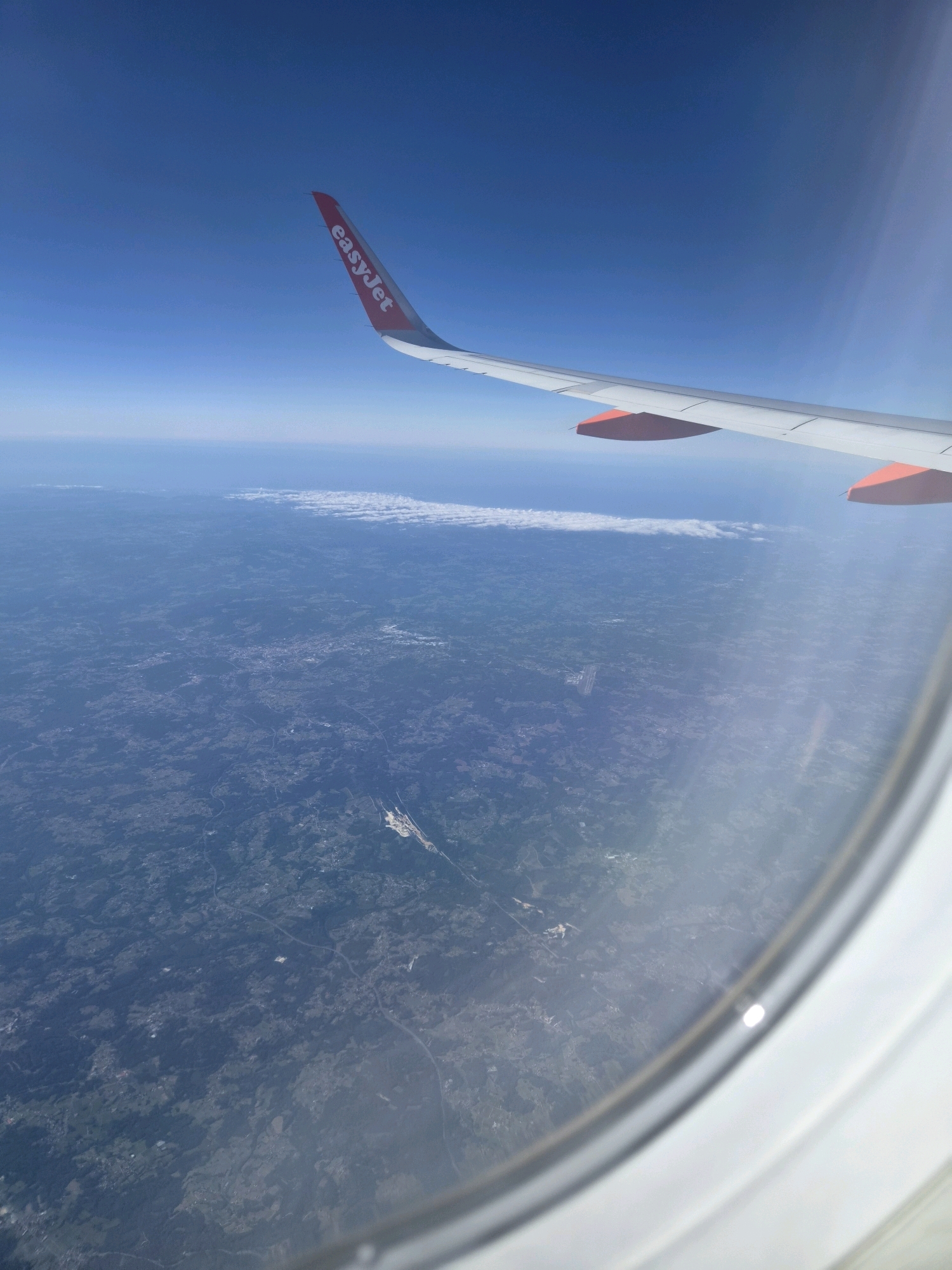 An aerial view from an airplane window shows the landscape below, with the wing of the plane visible in the upper portion of the image. The wing is white with orange accents at the tips and displays the "easyJet" logo. Below the plane, a vast expanse of land is visible, characterized by a mosaic of green fields, wooded areas, and scattered settlements. A thin band of clouds stretches across the horizon. In the foreground, the curved frame of the airplane window is visible. While no specific landmarks are immediately identifiable for Bazeilles-sur-Othain, the image depicts a rural or semi-rural area from a high altitude.