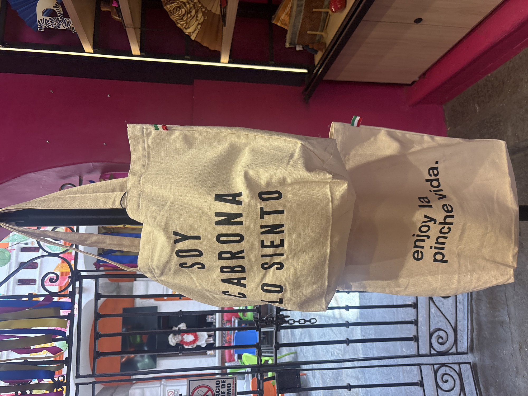Two cloth shopping bags hang from a wrought-iron gate in Puebla, Mexico. The bag on the left reads "Soy Cabrona Lo Siento" in bold black lettering, and the bag on the right reads "enjoy la pinche vida." The gate has colorful streamers tied to it and opens to an interior space with chairs and decorations. A bright pink wall is visible in the background, with shelves displaying various decorative items.