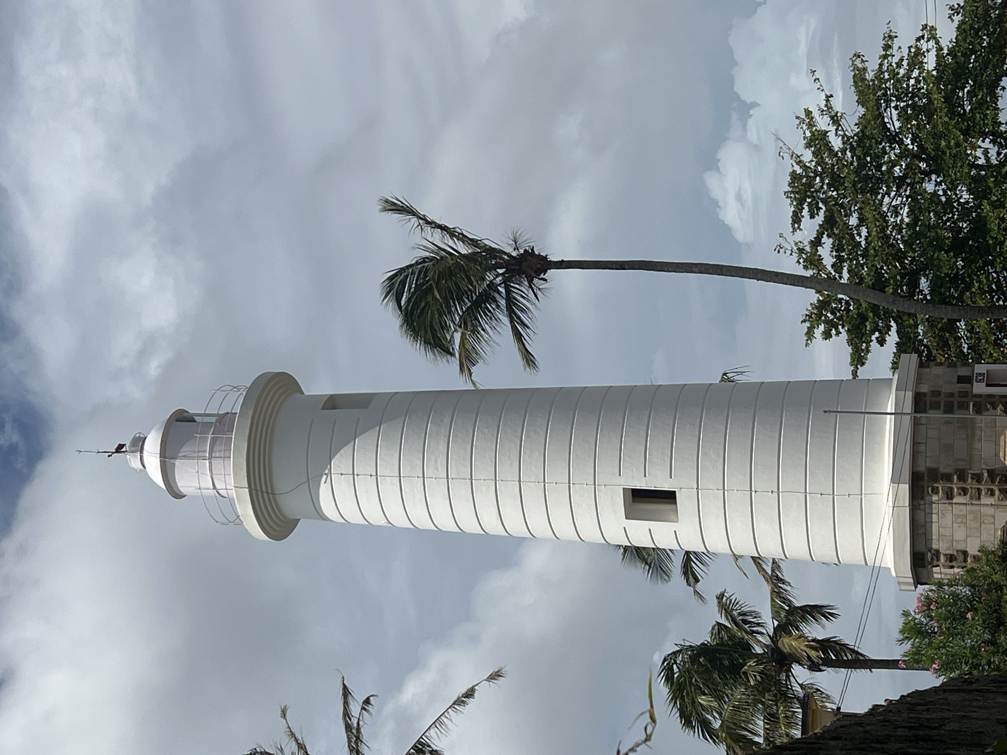 A tall, white lighthouse stands prominently against a cloudy sky in Galle, Sri Lanka. The lighthouse has a cylindrical body with horizontal ridges and features a small, square window on its lower section. At the top, it has a lantern room with a railing and a pointed roof. Palm trees with fronds blowing in the wind are visible on either side of the lighthouse, as well as some greenery and a stone base. The overall impression is of a coastal scene with weathered elements under a dramatic sky.