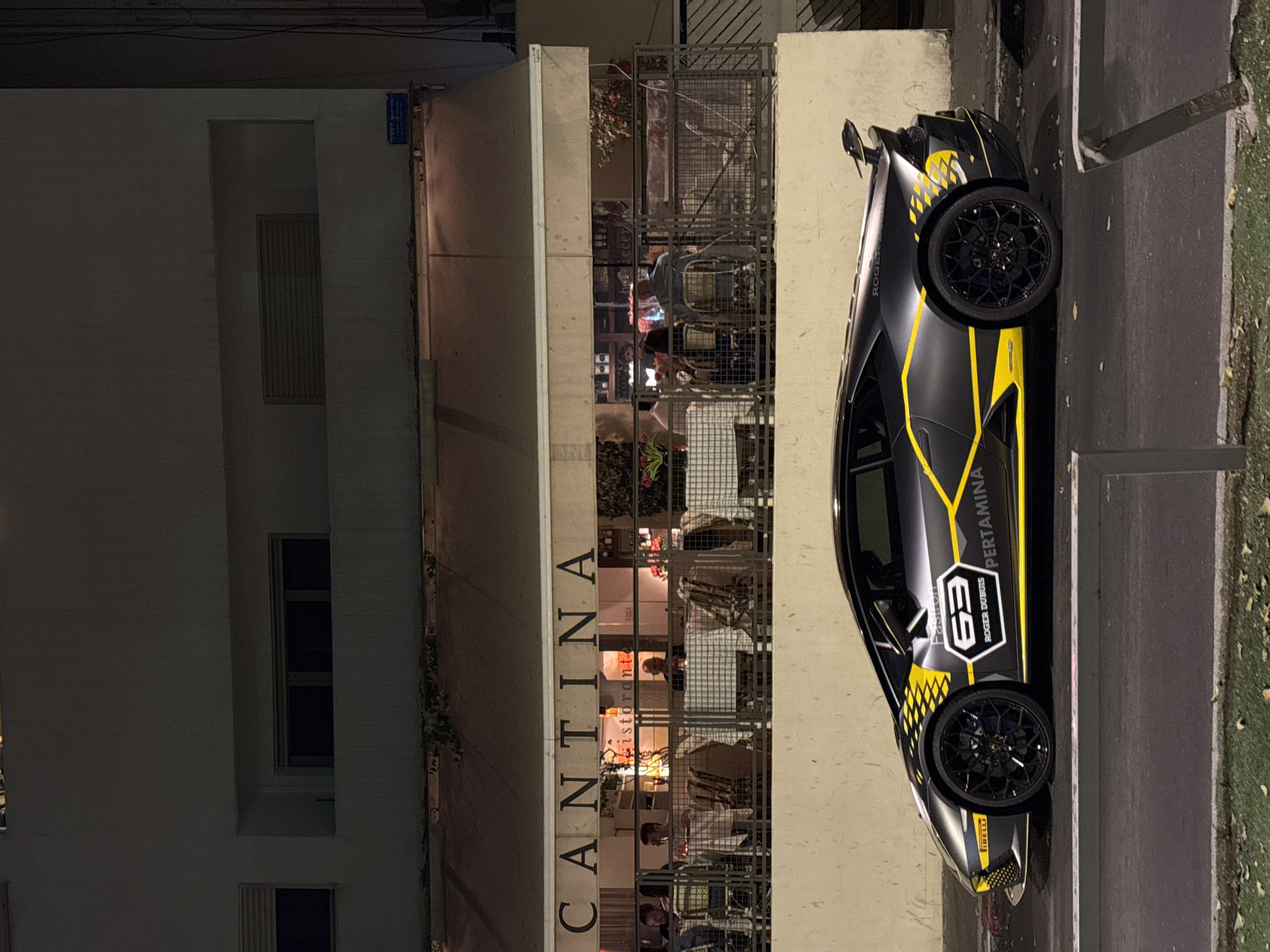 This nighttime scene depicts a black and yellow sports car parked alongside a building that houses a restaurant in Tel-Aviv, Israel.
The main subject is a high-performance sports car, likely a Lamborghini, customized with a matte black body and striking yellow accents. These include bold stripes, a checkerboard pattern, and various sponsor decals. Visible text on the car's side includes "PERTAMINA," "ROGER DUBOUIS," "63," "IRELLI," and partially "ROGUE" and "FAST-R." The car features intricate black wheels with yellow details, and the "PIRELLI" brand name is visible on its tires. It is parked on a dark paved surface next to a narrow strip of grass and a curb.
To the left of the car, a multi-story building features a brightly lit restaurant or café on its lower level. The restaurant's name, "CANTINA," is displayed in large, dark letters on the building's light-colored facade. Through large windows or an open-air design, several people are visible inside, seated at tables and engaged in dining or socializing. Smaller text within the restaurant reads "ristorante." The interior is warmly lit, casting light onto the street. Some plants are visible near the restaurant's entrance and on the ground beside the car.
The upper floors of the building appear dark, with various windows. A blue street sign with Hebrew text, "רחוב וולפסון" (Wolfson Street), is affixed to the building's upper left section, confirming the Tel-Aviv location. The overall setting is an urban environment illuminated by artificial light, characteristic of an evening or nighttime scene.
