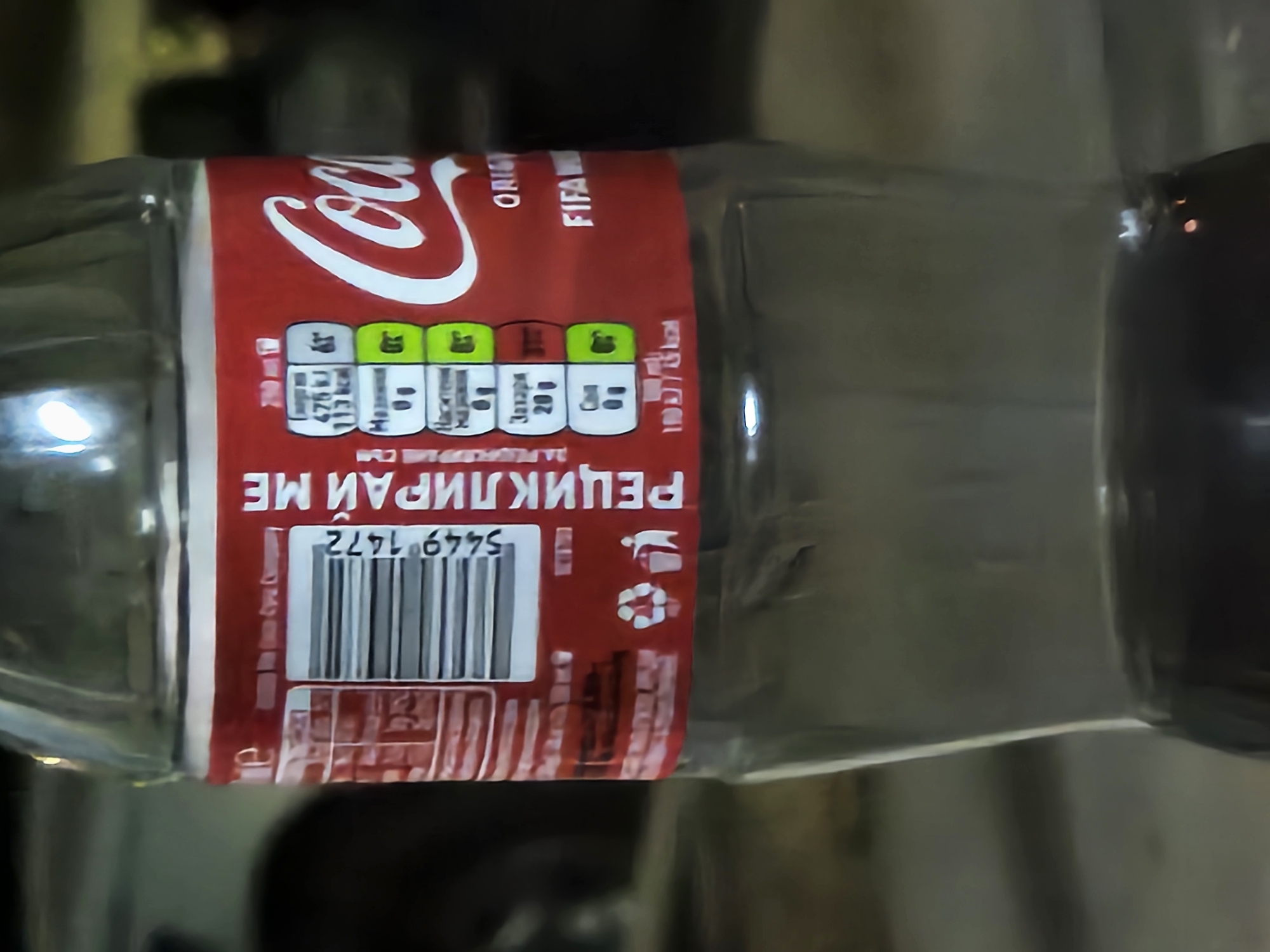 A close-up, low-angle shot shows the label of a clear plastic bottle of Coca-Cola. The label is red with white text and graphics. The Coca-Cola logo is prominently displayed on the right side of the label, in white cursive font. To the left of the logo, there is a nutritional information panel with values for energy, fat, and sugar, presented with green and white icons. A barcode is also visible on the left side of the label. Below the nutritional information, there are recycling symbols and a trash bin icon. The bottle appears to be partially filled with a dark liquid. The background is out of focus and appears to be an indoor or outdoor setting with muted colors. There is no specific indication of the city of Biaga, Bulgaria in the image.