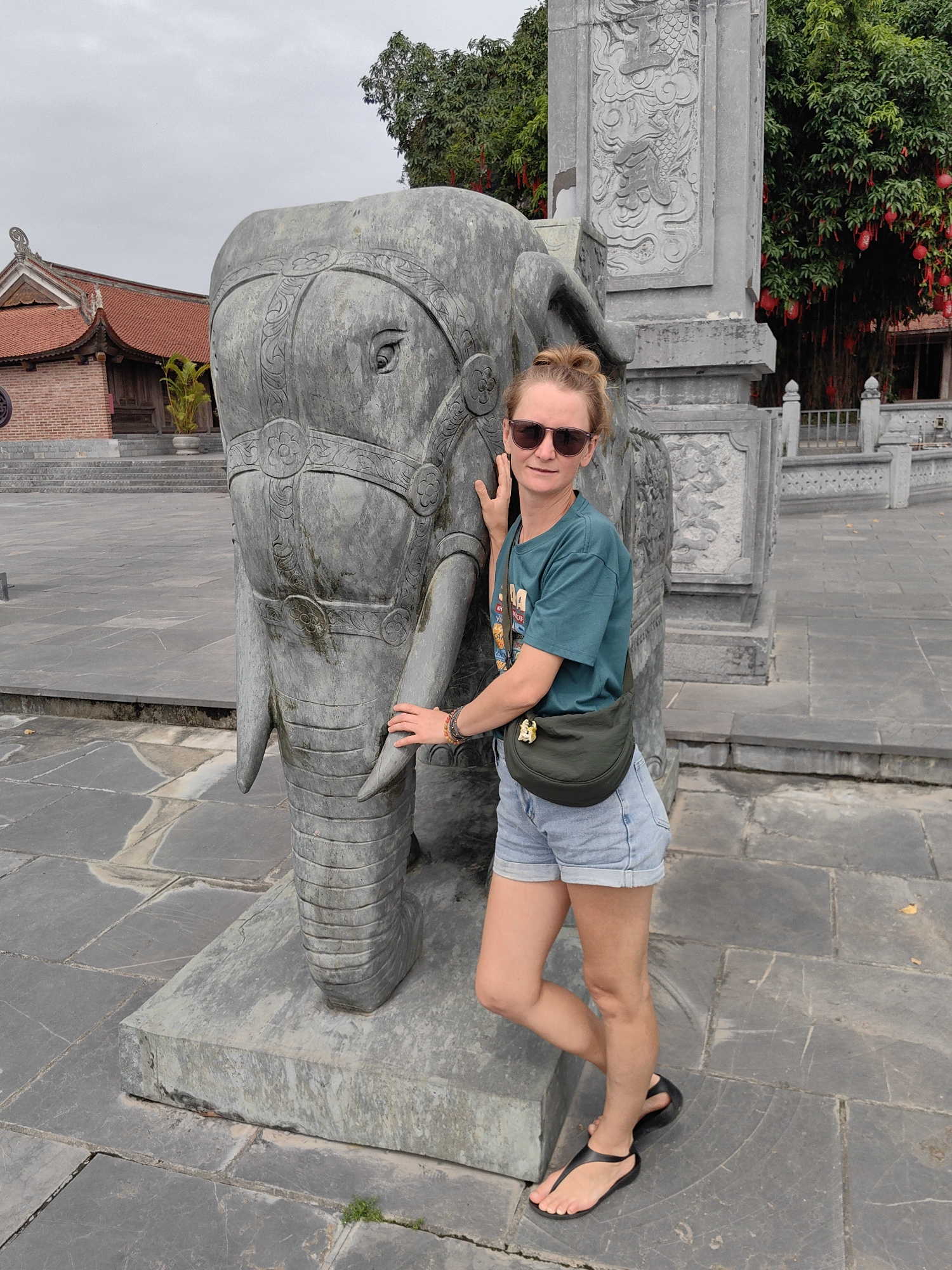 A woman poses next to a large, intricately carved grey stone elephant statue at a historical or cultural site in Hong Gai Ward, Vietnam. The woman has blonde hair styled in a bun and wears dark sunglasses, a teal t-shirt, light denim shorts, and black flip-flops. A dark green fanny pack is slung across her body. She stands with a neutral expression, looking towards the camera, with her right hand resting on the elephant's head and her left hand near its trunk, leaning slightly against the statue.
The stone elephant statue is adorned with decorative patterns carved into its head, ears, and body, and stands on a rectangular stone base. The ground is paved with large, grey flagstones. In the background, traditional Vietnamese architecture with distinctive red-tiled roofs and brick walls is visible, suggesting a temple or pagoda complex. To the right, a tall, elaborately carved stone pillar stands, and behind it, a tree is festooned with numerous red lanterns, adding a festive cultural element. The sky is overcast, indicating a daytime photograph taken under soft, diffused light. While some text is visible on the woman's t-shirt and carved into the stone pillars, it is not clearly legible.