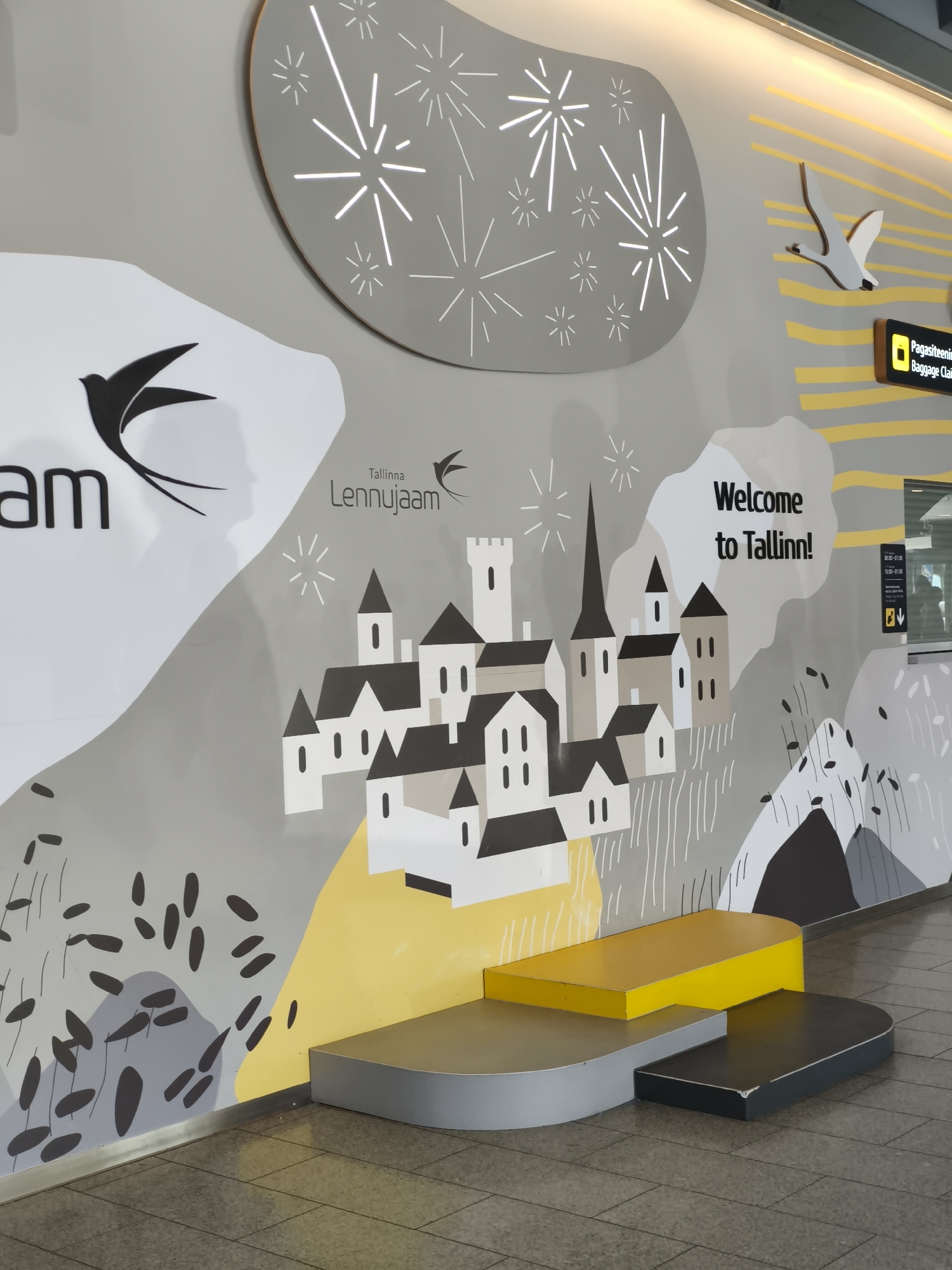 The image shows a modern art installation on a wall, likely at an airport or public transportation hub in Tallinn, Estonia, given the "Welcome to Tallinn!" text. The artwork features stylized drawings of fireworks, a swan, a flock of birds, and a simplified cityscape with towers and houses. A curved bench with tiered seating in yellow, gray, and black is positioned in front of the wall. The floor is tiled. To the right of the artwork, there is a sign indicating "Baggage Claim." The overall aesthetic is contemporary and minimalist, with a color palette dominated by gray, white, yellow, and black.
