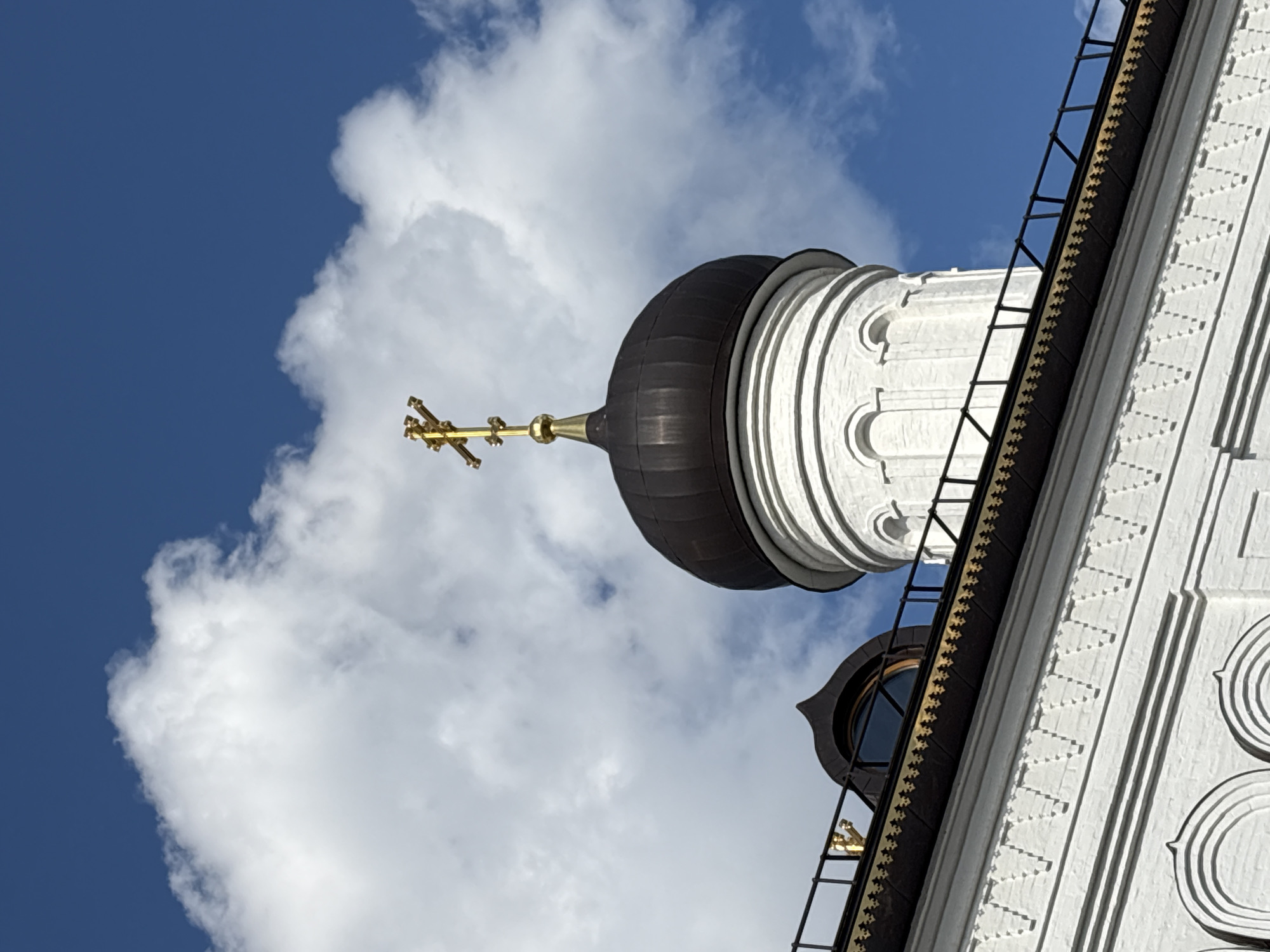 The image, taken from a low angle, features a section of a white, intricately patterned building with a black dome and a golden cross atop it. The structure is set against a backdrop of a partly cloudy blue sky. A ladder runs up the side of the building. The location is Ulyanovsk, Russia, based on the likely architectural style of the church.