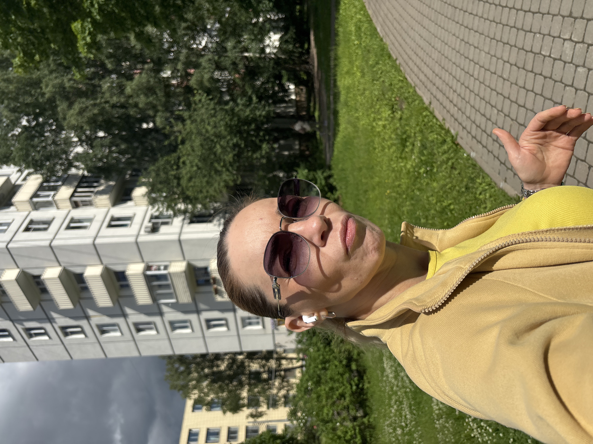 The image is a close-up, selfie-style shot of a woman in Saint Petersburg, Russia. She is outdoors, walking on a paved path next to a grassy area with small white flowers. In the background, there is a tall, grey, multi-story apartment building with balconies, and lush green trees. The woman is wearing large, dark sunglasses, a yellow zip-up jacket over a yellow shirt, and white wireless earbuds. Her hair is pulled back. Her left hand is visible, with fingers slightly curled. The lighting suggests it is daytime, with sunlight casting shadows. The overall atmosphere is casual and urban.