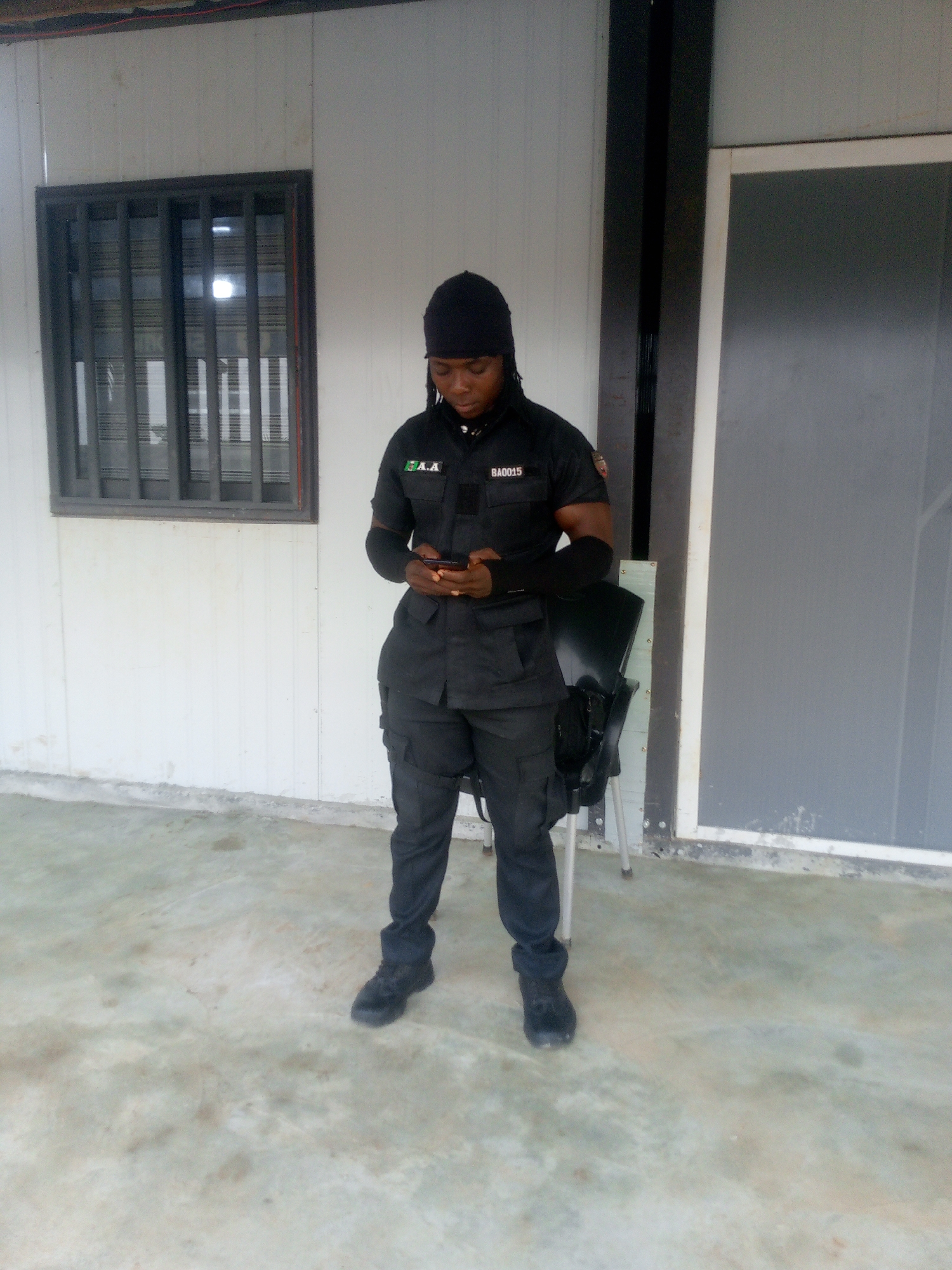 A person wearing a black uniform, including a black cap, is standing outdoors, looking at a smartphone. The uniform has patches on the sleeves, one of which reads "BA0015". The person is wearing black trousers and black boots. They are standing on a concrete surface in front of a white building with a window on the left and a door on the right. A black chair is to the right of the person. The scene appears to be in Indo, Nigeria.
