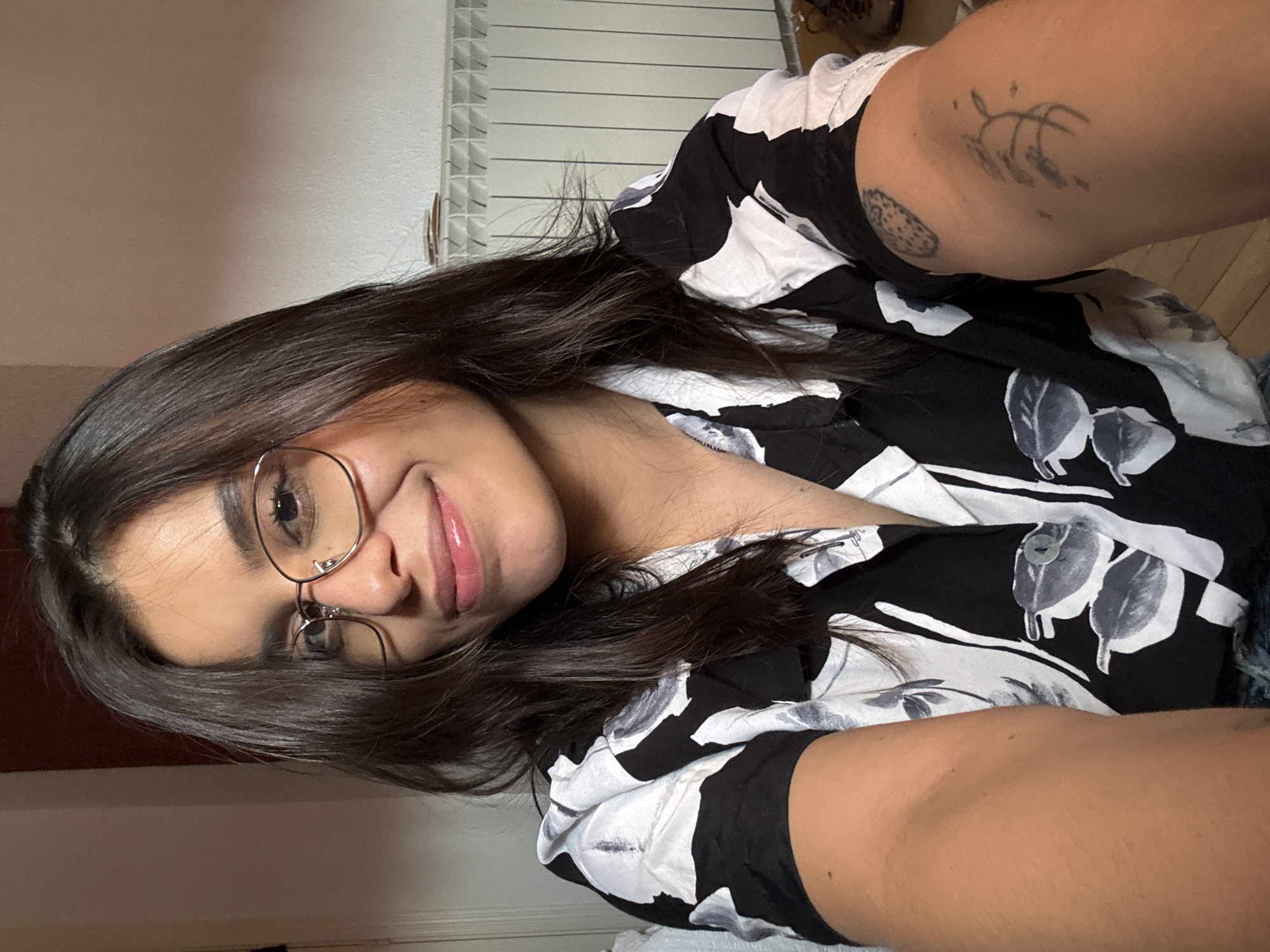 A young woman with dark hair and glasses is shown in a close-up selfie. She is wearing a black and white patterned shirt. Her left arm is extended towards the camera, and her right arm is visible on the right side of the frame, featuring a tattoo. The background includes a radiator and a wall. The image does not provide any information that allows for its location to be specified in the city of Las Rozas de Madrid, Spain.