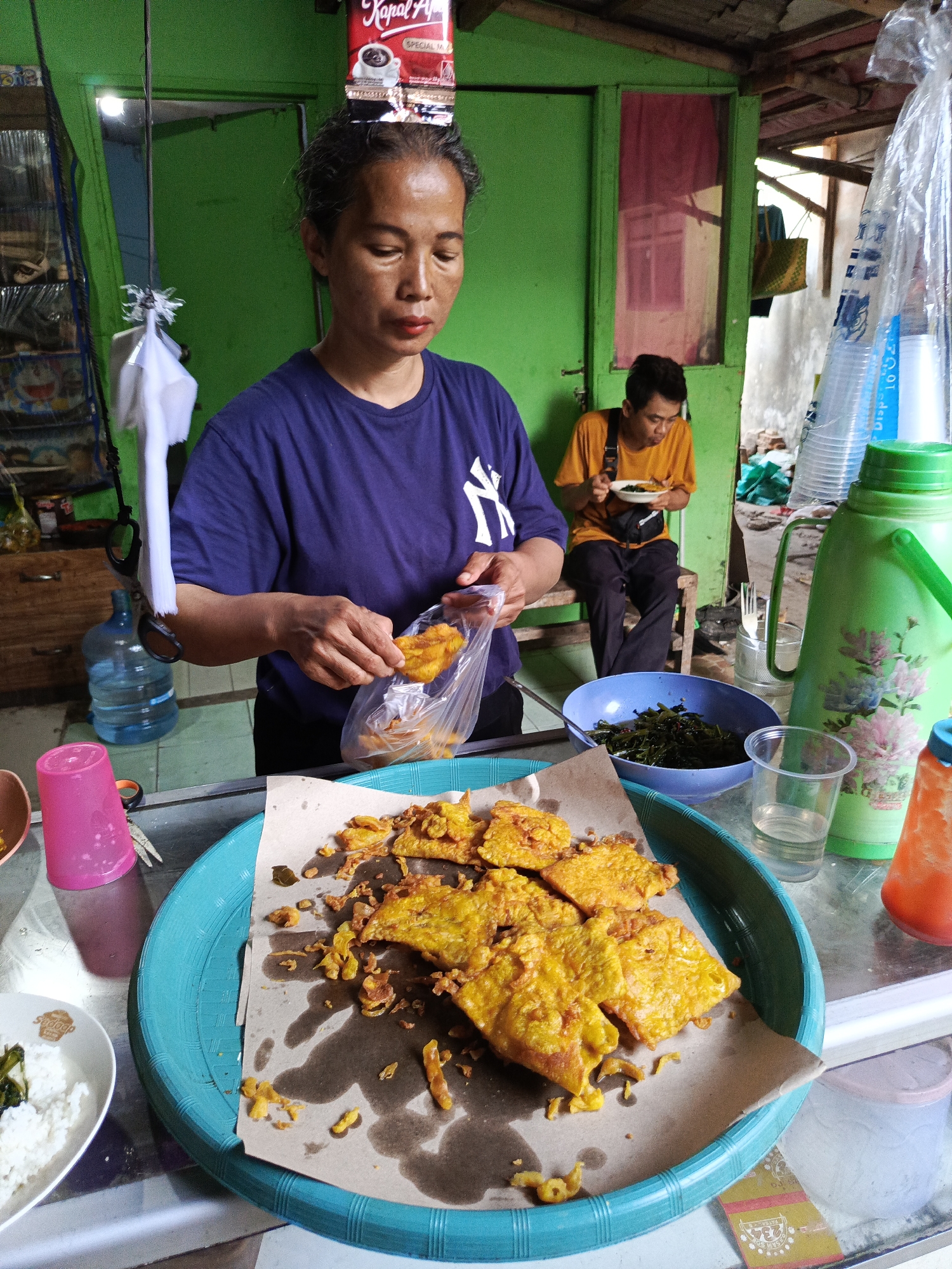 The image depicts an outdoor food stall in Tangerang, Indonesia. A woman in a purple t-shirt is preparing fried food, likely tempeh or a similar batter-coated item, holding a piece in a plastic bag. In the background, a man in a yellow t-shirt is seated and eating from a plate. Various food items and serving dishes are visible, including a large turquoise platter filled with fried food, a blue bowl of green vegetables, a white bowl of rice, and a pink cup. A large green thermos is also present, along with stacks of disposable cups. The environment suggests a humble, everyday setting, possibly a street food vendor or a small eatery. The backdrop shows a green painted wall and a wooden structure, indicative of an open-air establishment.
