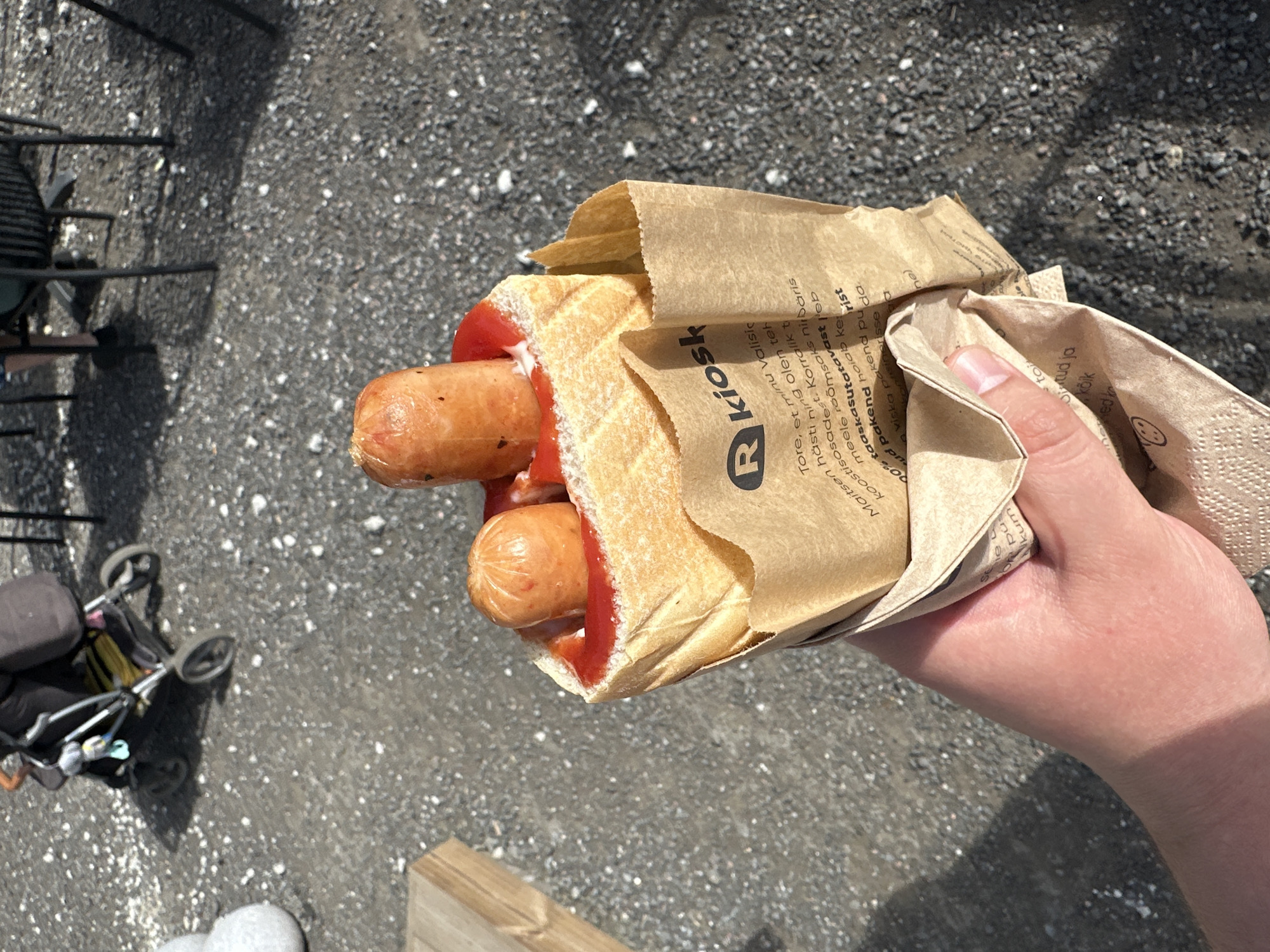 A hand holds a large hot dog in a paper wrapper, with two sausages on top and covered in ketchup, placed in front of a blurry background. The wrapper has text in a foreign language that appears to be in Estonian, with a logo that reads "R kiosk". The background shows a stroller, a table with chairs, and a grey gravelly ground, suggesting an outdoor urban setting.