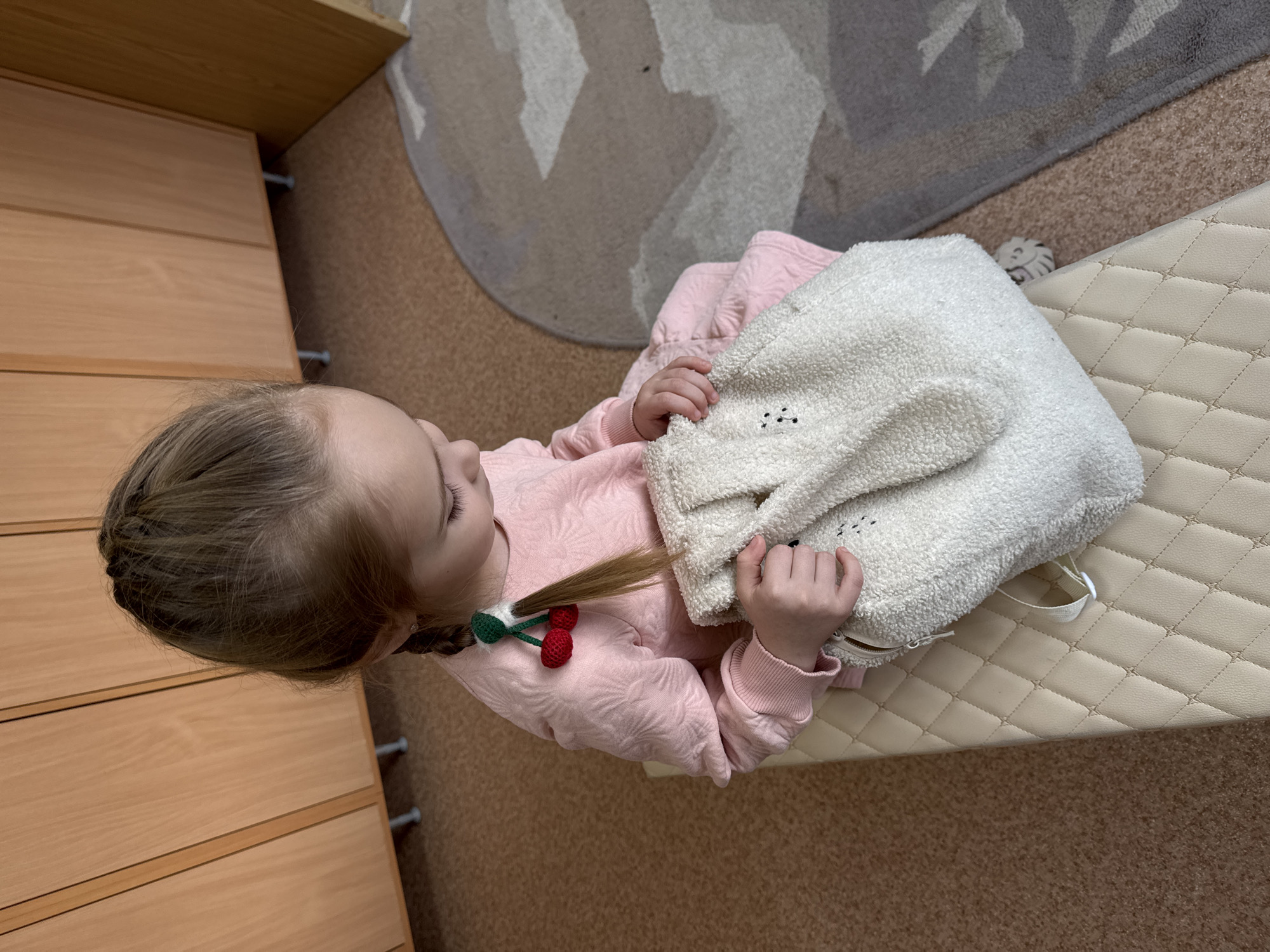 In the city of Chaikovsky, Russia, a young female child is seated on a cream-colored, quilted bench in an indoor setting. She has light brown hair styled in a braid, which is adorned with a red and green cherry-shaped accessory. The child wears a light pink, textured long-sleeved top and is looking down, holding a white, fuzzy backpack with both hands. The backpack features an animal design, possibly bunny ears, and small black stitched details resembling a face. Behind the child, light-toned wooden cabinetry with multiple vertical panels stands on silver support legs. The floor transitions from a light brown carpet in the foreground to a grey carpet with a geometric pattern in the mid-ground.