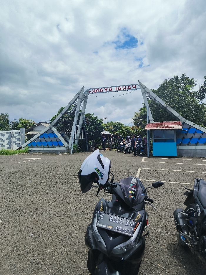 The image shows a black motorcycle parked in front of an entrance gate. The gate is made of metal and is shaped like a triangle with a sign that reads "PARAYANAG" in reverse. There are people standing behind the gate. There are several other motorcycles parked behind the gate. There is a blue and white striped wall to the left of the gate and a blue building to the right. The scene is in the town of Majalengka, Indonesia, and the coordinates are -6.846, 108.204. The weather is cloudy and there is a blue sky in the background. The foreground is a parking lot. The motorcycle has a white helmet on it. The motorcycle has a license plate that reads "E 6345 XS". The motorcycle has a sticker on it that says "YAMAHA".