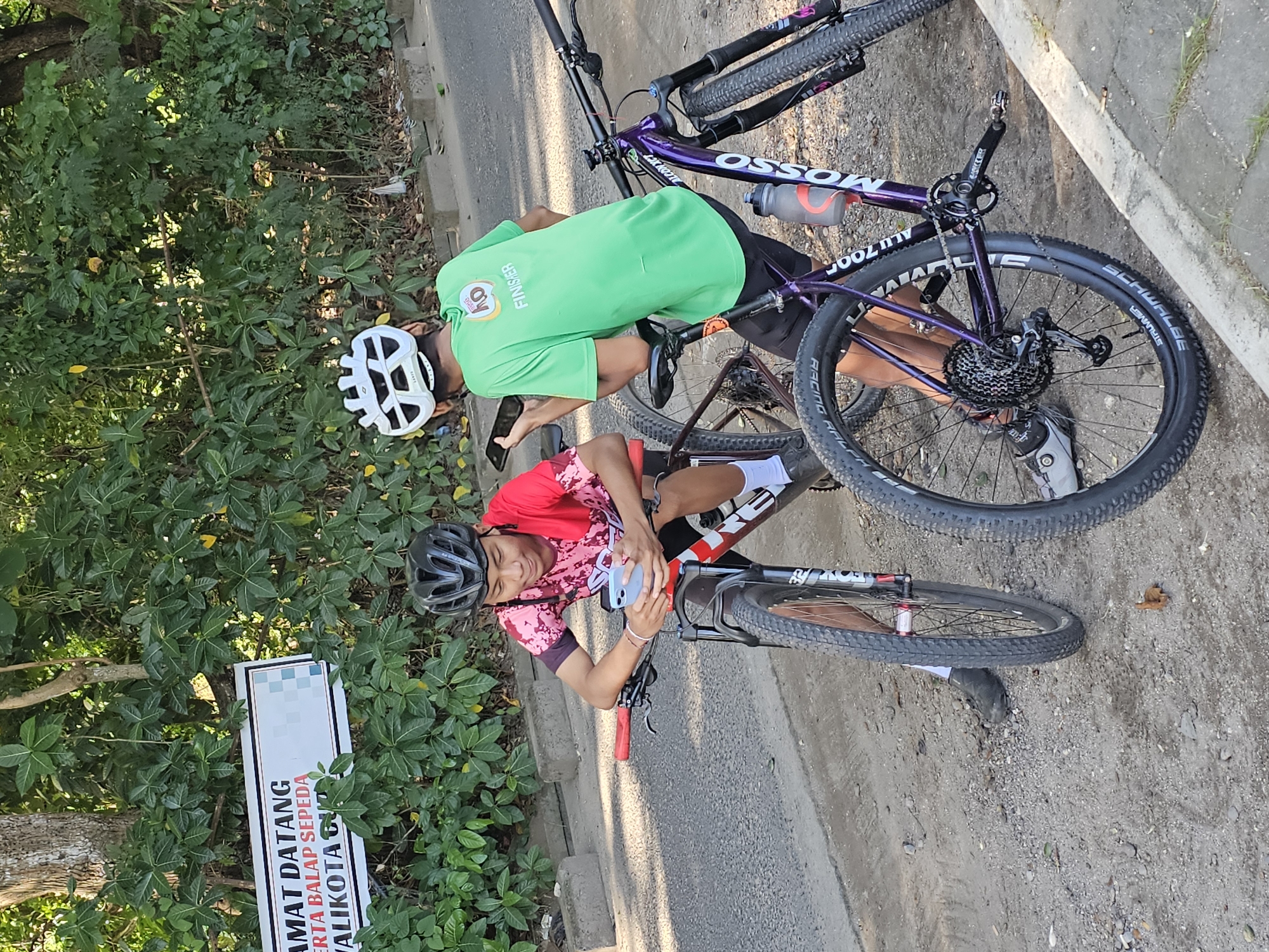 Two cyclists stand with their bikes on a road in Denpasar, Indonesia, indicated by the sign "SELAMAT DATANG SERTA BALAP SEPEDA BALIKOTA DENPASAR." One cyclist, wearing a red and pink shirt and a black helmet, looks at a blue smartphone held in their hands. The second cyclist wears a green shirt, black shorts, a white helmet, and black shoes and is also using a phone. Both bikes are mountain bikes; one is purple and the other is red and white. The environment includes foliage and the edge of a paved road or sidewalk.