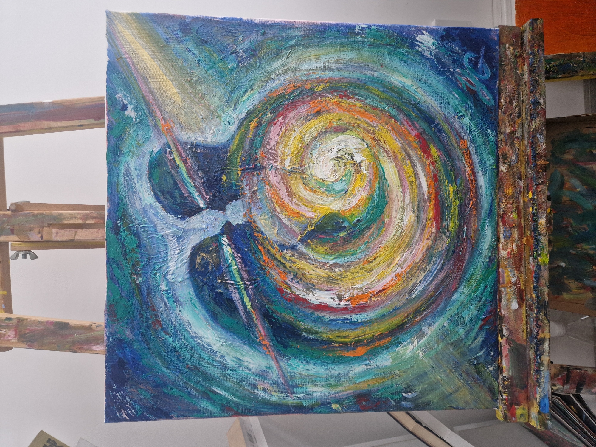 The image shows a square-shaped canvas painting resting on an easel. The painting features an abstract design with a swirling pattern of colors, predominantly blues, greens, yellows, oranges, and hints of red. The brushstrokes are thick and textured, creating a dynamic and vibrant effect. The central part of the swirl is a mix of white, yellow, and pink. In the upper left corner, there is a ray of light. The painting is leaning against the easel, which is stained with paint from previous use. The background includes a white wall and parts of other easels and canvases. The location appears to be an artist's studio, city of Unknown, Unknown.