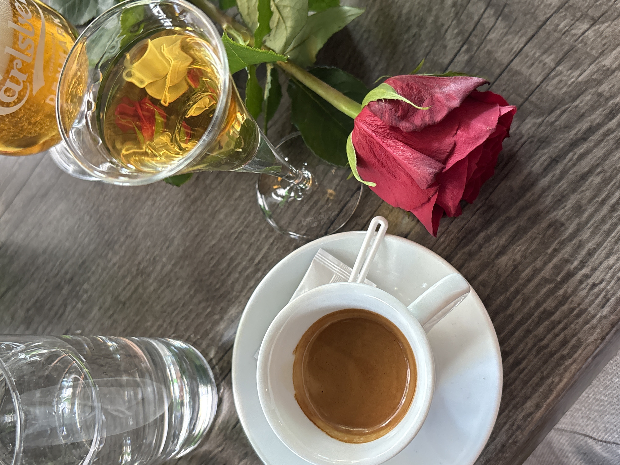 An overhead, flat lay shot captures a collection of drinks and a single red rose arranged on a rustic wooden table. The rich grain of the wooden surface runs diagonally, providing a textured backdrop for the items.
Dominating the upper section of the image is a vibrant red rose, its petals showing slight variations in texture, and its stem and green leaves extending towards the left. Adjacent to the rose, a champagne flute holds an amber-colored liquid, possibly a sparkling wine or cider. Intricate reflections of its surroundings, including what appears to be part of a yellow object and some red elements, are visible within the liquid. Partially cropped on the far left, a bottle prominently displays the "Carlsberg" logo and the word "DENMARK," confirming it as a beer.
In the lower right portion of the frame, a white espresso cup filled with a rich, dark brown espresso shot, topped with a creamy layer of crema, sits centered on a matching white saucer. Two neatly packaged sugar packets and a white stirrer are also placed on the saucer. To the left, a clear, empty glass of water adds to the array of beverages.
The scene suggests a relaxed atmosphere, likely in a cafe or restaurant in Kragujevac, Serbia. The natural lighting indicates it is daytime. The presence of the rose could imply a special occasion or a romantic encounter. The overall impression is one of calm enjoyment and leisure. No people are visible in the image.