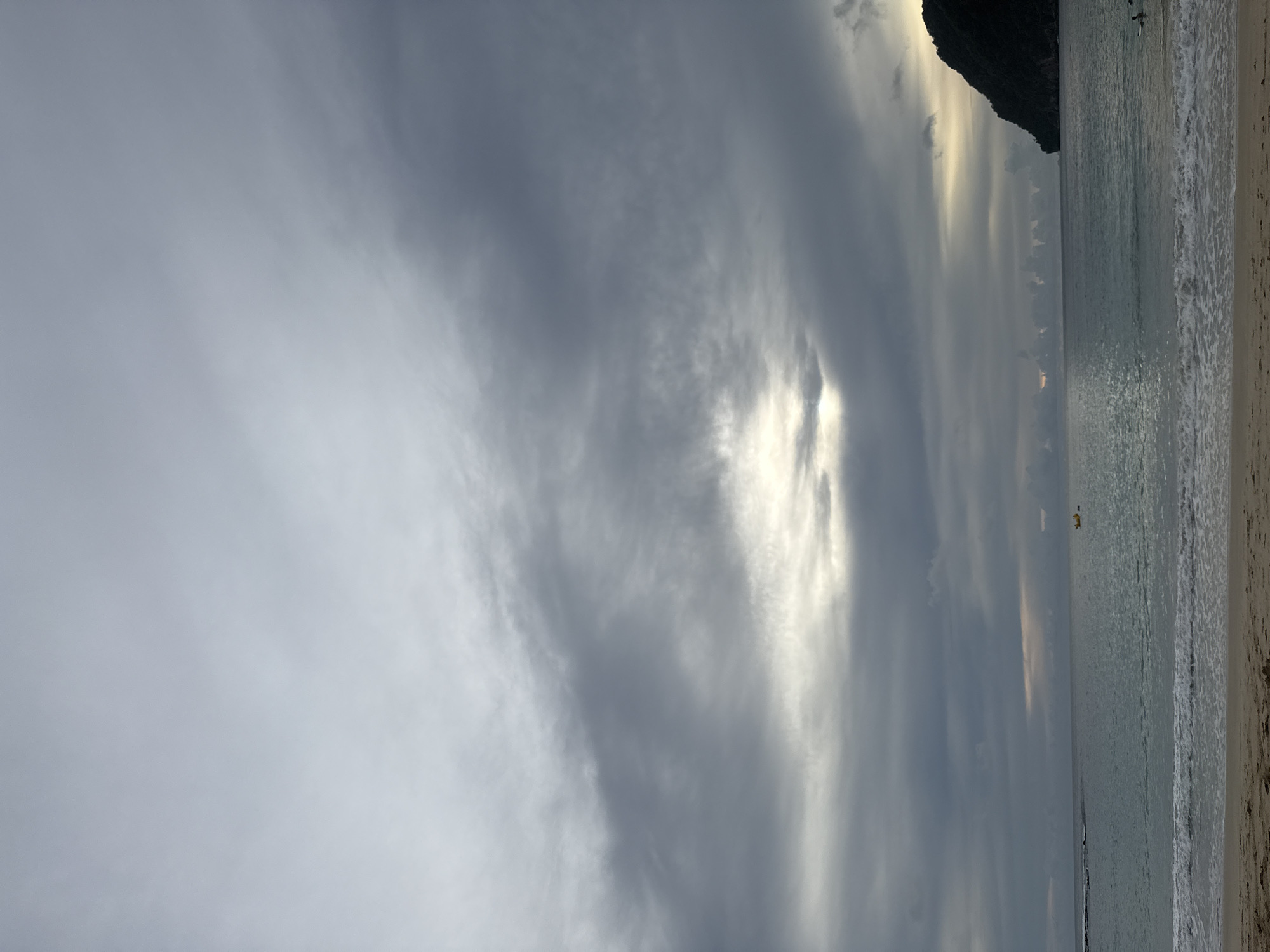 This is a wide-angle, eye-level shot of a beach scene on a cloudy day, likely in Meunasahbale, Indonesia. The sky is filled with heavy, gray clouds, with a bright patch of sunlight breaking through in the middle distance. The sea is calm with gentle waves lapping at the sandy shore. In the water, a small yellow boat is visible in the distance. To the right, a dark, tree-covered hill juts out into the sea. There are two people visible in the water near the shore on the right side of the frame, with one appearing to be a swimmer. The sand in the foreground is a light brown color, with some subtle ripples from the receding tide.