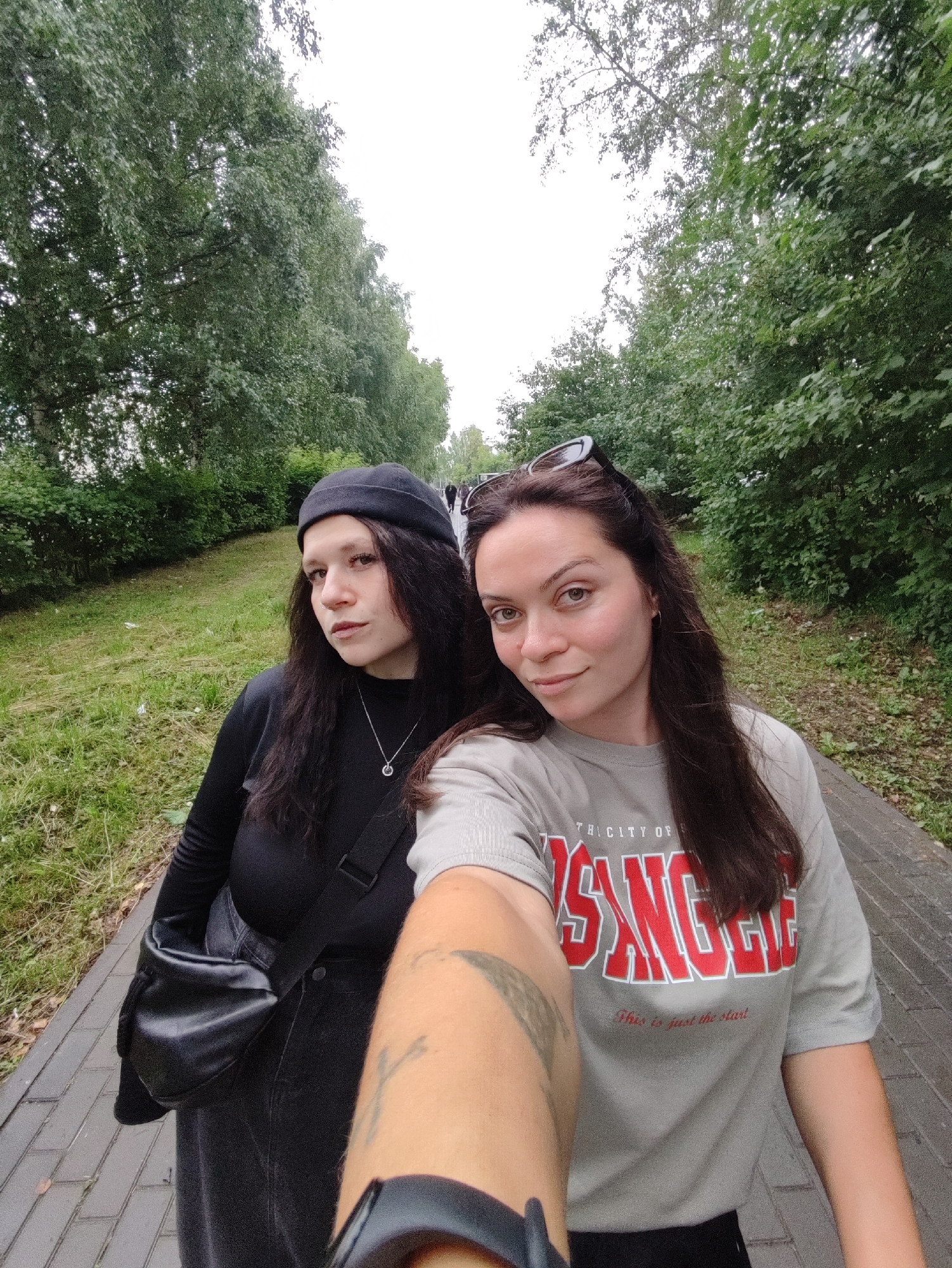The image shows two young women posing for a selfie outdoors. The woman on the left is wearing a black beanie, a black long-sleeved shirt, and a black crossbody bag. She has long, dark hair and is looking towards the camera with a neutral expression. The woman on the right has long, dark hair and is wearing a light gray t-shirt with red lettering. She is smiling slightly and looking at the camera. Sunglasses are perched on her head. Their arms are in the foreground, indicating a selfie is being taken.
They are standing on a paved walkway lined with grass and trees. The trees are lush and green, suggesting a warm weather season, possibly summer. The sky appears overcast. In the background, more trees and foliage are visible, and a few other people can be seen walking further down the path, suggesting a public park or walking trail.
The t-shirt of the woman on the right has visible text: "THE CITY OF" and "LOS ANGELES" in large red block letters. Below that, smaller text reads, "This is just the start".
The overall impression is a casual outdoor activity, perhaps a walk or outing. The emotions conveyed are relaxed and friendly. The location context provided indicates Chaikovsky, Russia, but the visible text on the t-shirt references Los Angeles, creating a contrast.