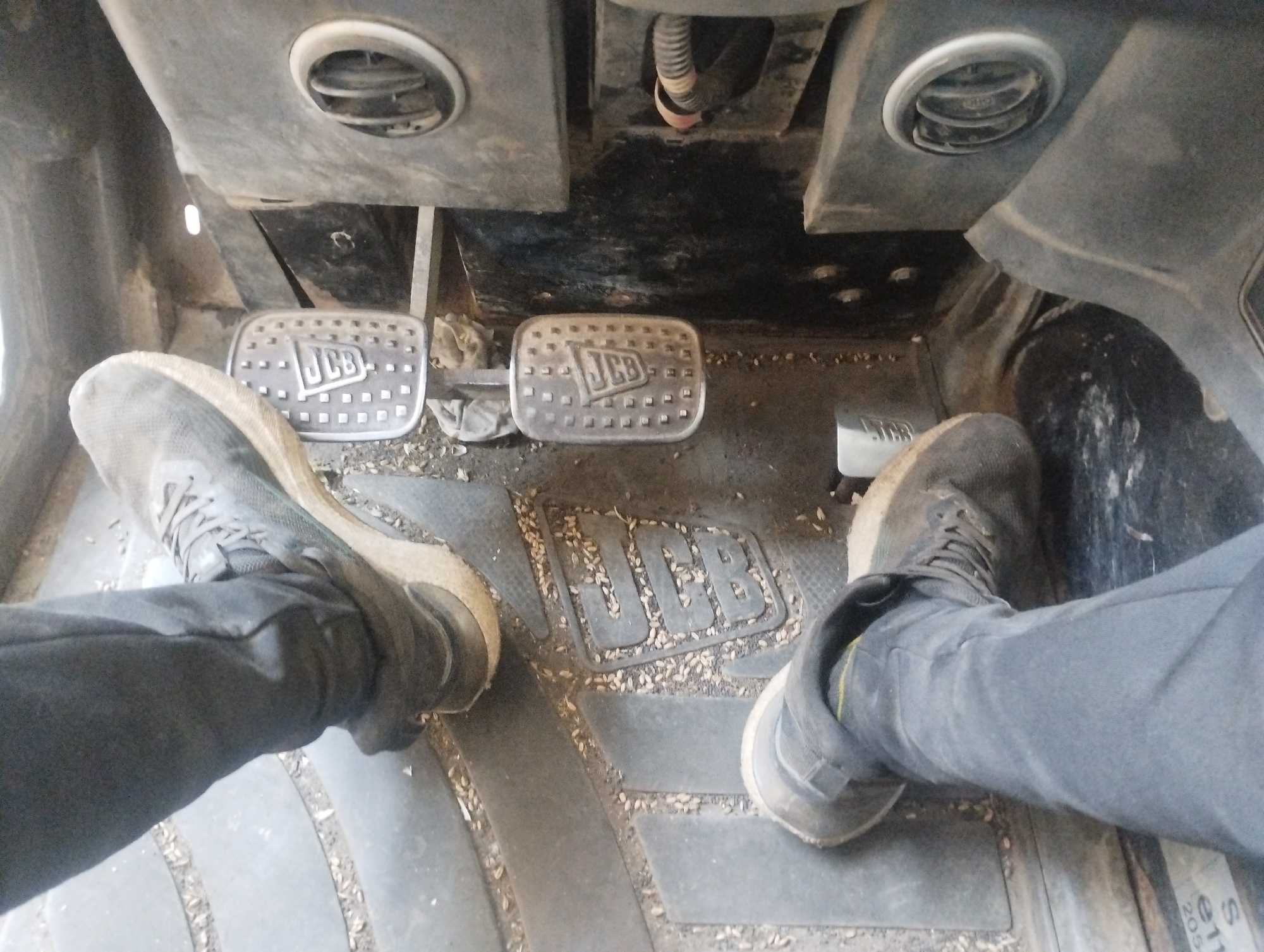 Two pairs of feet in dark trousers and athletic shoes are visible within the driver's cab of a vehicle, likely a piece of heavy machinery given the presence of two distinct pedals bearing the "JCB" logo. The pedals are metallic and textured for grip. The floor of the cab is covered by a dark rubber mat with a raised pattern, speckled with small, light-colored debris, possibly grain. Two circular air vents are positioned above the pedals. The overall environment appears utilitarian and somewhat worn. The image was captured in Sredets, Bulgaria.