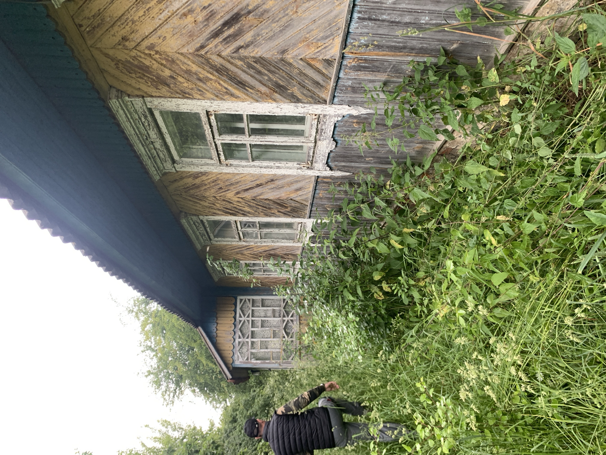 A man wearing a black vest over a camouflage long-sleeved shirt and grey pants walks away from the camera through tall green grass and foliage. He is approaching a weathered, old wooden house with peeling yellow paint and a dark blue, corrugated roof. The house has several windows, one of which is partially obscured by overgrown plants. To the left of the main house is a smaller, enclosed porch or extension with a decorative window. The scene appears to be in a rural or overgrown area, consistent with the location of Блеўчыцкі сельскі Савет, Belarus, given the architectural style and the natural surroundings. The sky is overcast.