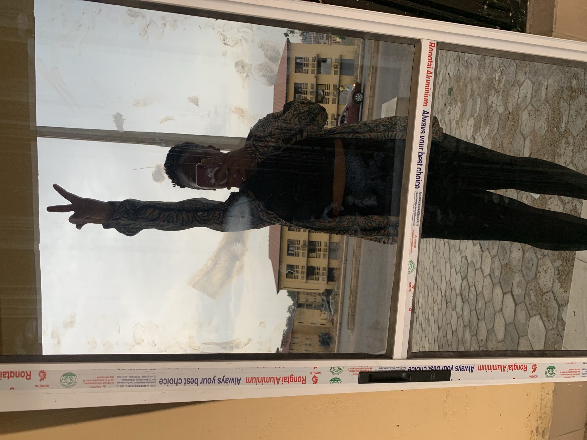 A reflection of a person in a window is visible, with the person holding up a peace sign. The window frames are labeled "Rongtai Aluminium," a company apparently located in Jos, Nigeria, as the label mentions "RONGTAI ALUMINIUM (NIGERIA) CO.LTD." The reflection shows a building across the street with several windows, along with a vehicle parked on the road. The window is slightly dirty with smudges. The exterior wall surrounding the window frame is light yellow. The overall environment appears to be urban with a residential or commercial building visible.