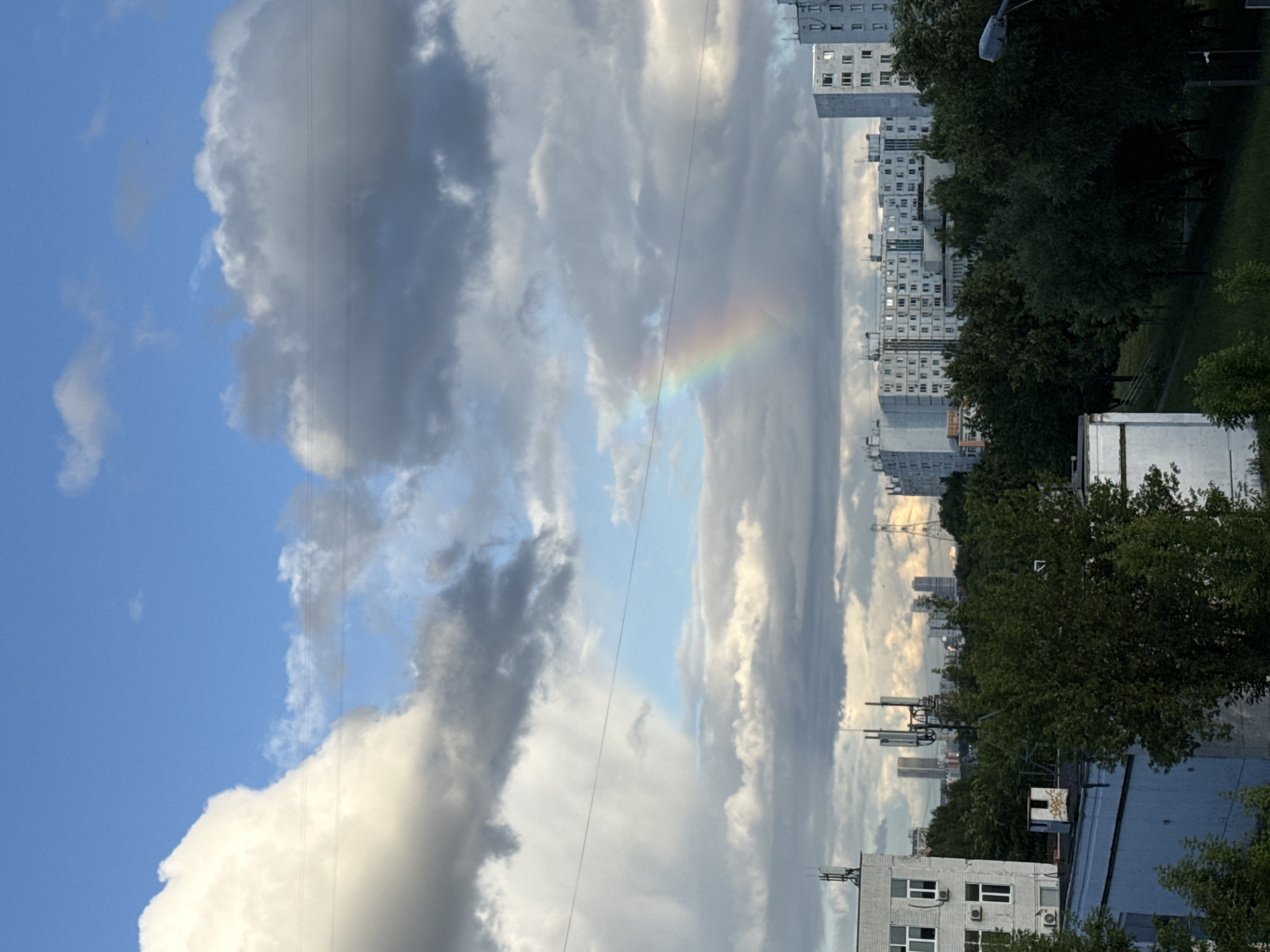The image captures a cloudy sky over a city landscape, with a faint rainbow visible in the upper center. The sky is a mix of bright blue and dark gray clouds, with a large, fluffy white cloud dominating the upper left quadrant. A thin wire stretches horizontally across the sky. Below the sky, a dense line of green trees separates the sky from the urban environment. In the background, a series of apartment buildings, some tall and white with multiple windows, are clustered together. A few taller structures, likely communication towers or other urban infrastructure, are interspersed among the buildings. The foreground features the roofs of two lower-rise structures, one white and one light blue, with some pipes and antennas visible. The overall impression is an urban scene with a natural phenomenon occurring in the sky. The architecture suggests a location in Moscow, Russia.