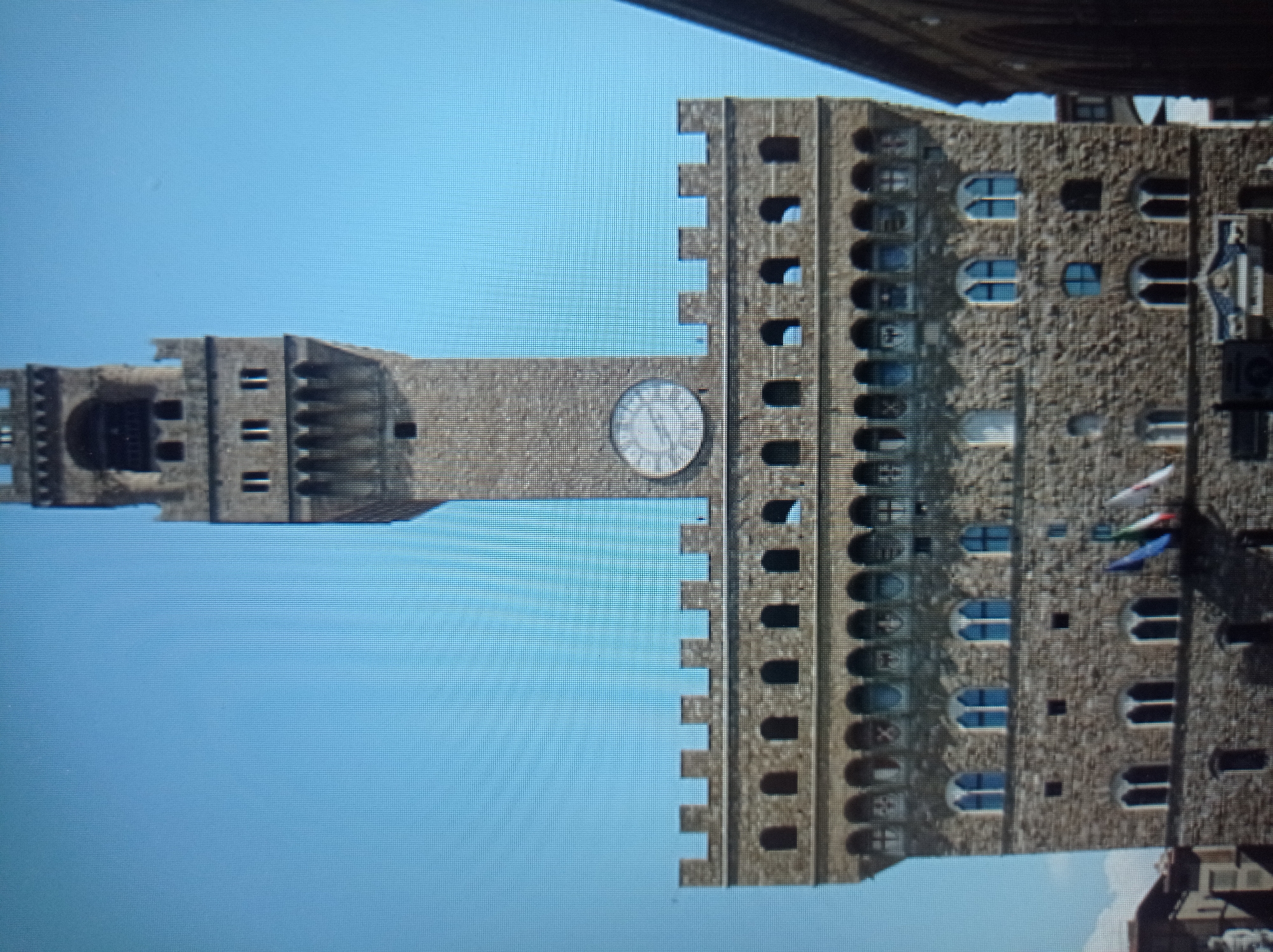 This image depicts the Palazzo Vecchio in Florence, Italy, under a clear blue sky. The imposing stone tower, a prominent feature of the building, dominates the center of the frame. It is adorned with a large clock face and features crenellations at its summit. The lower section of the building exhibits a series of arched windows and decorative friezes. Several flags, partially blurred, are visible near the base of the tower. The overall scene suggests an architectural landmark in an urban setting. There is no indication of Mukachevo, Ukraine, in this image.