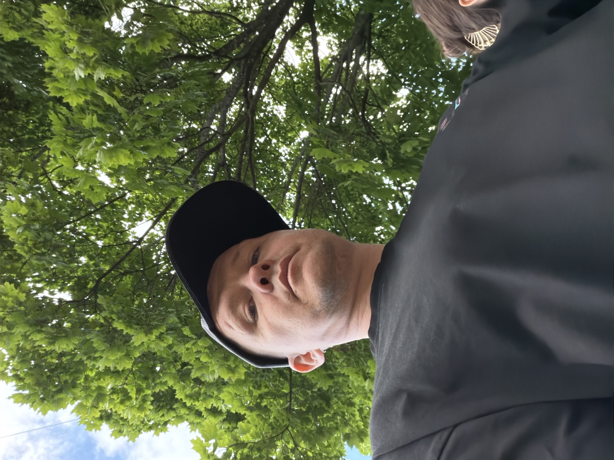The image features a low-angle shot of a person wearing a black baseball cap and a black jacket, looking up at the camera. The person's face is partially visible, with a light complexion and short hair. Their expression is neutral. To the left of the frame, lush green leaves of a tree are prominent, with branches extending across the upper portion of the image. The sky, a mix of blue and white clouds, is visible through the foliage. A hint of another person wearing a gold, fan-shaped earring and a dark top is visible in the upper right corner. The setting appears to be outdoors, likely a park or a wooded area, under daylight. The orientation of the image is rotated 90 degrees clockwise. There is no visible text in the image.