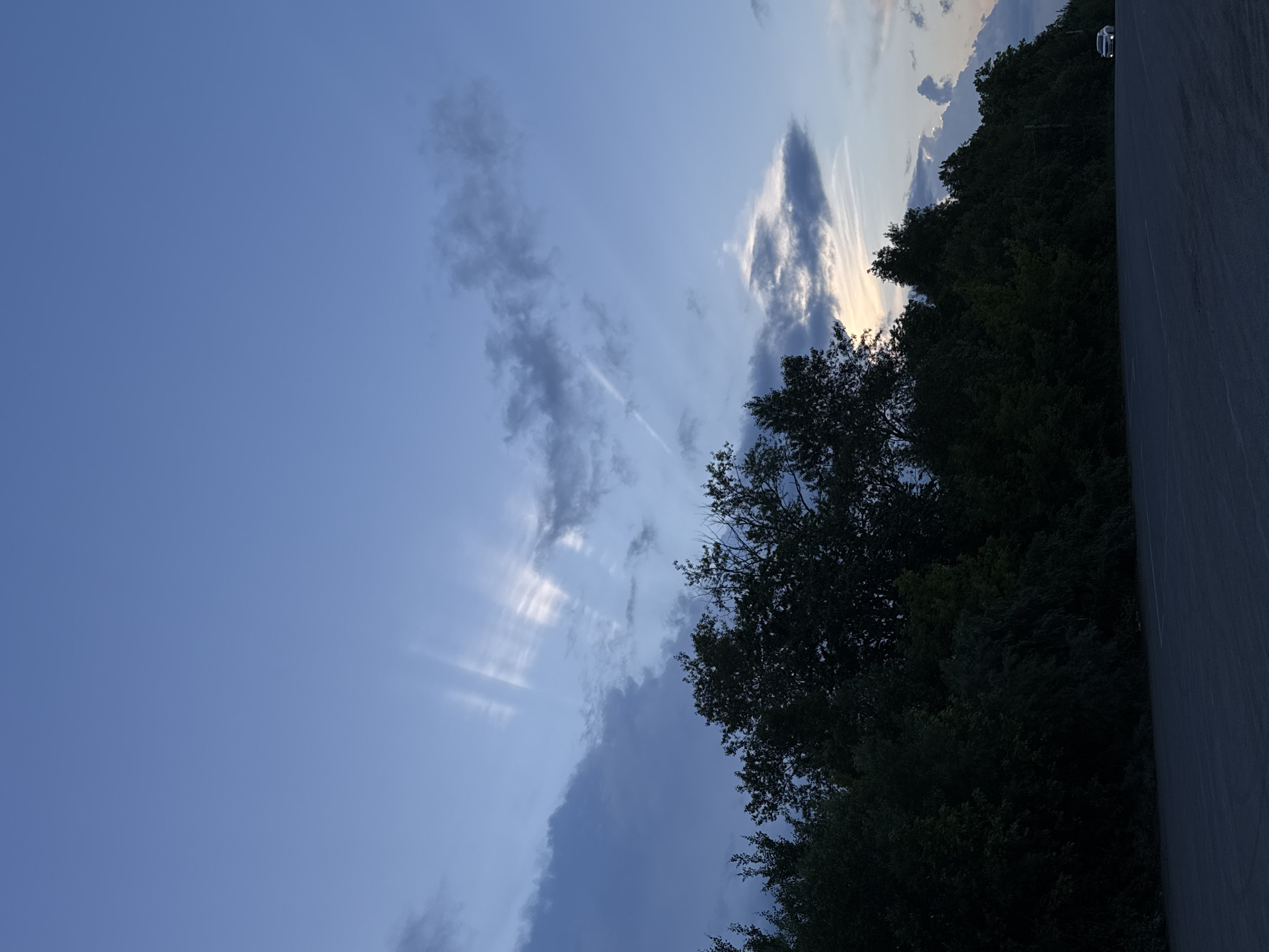 This image depicts a wide, empty paved area, possibly a parking lot or road, in the foreground. A dense line of dark green trees forms a thick barrier along the middle ground, obscuring much of what lies beyond. Above the trees, the sky is a gradient of deep blue to lighter shades of blue, with scattered clouds. Dramatic sunbeams radiate from behind some of the clouds, creating a dramatic effect. In the far right background, a single white car with its headlights on is visible, suggesting movement or arrival. The overall atmosphere is one of twilight or early evening. The image does not contain any specific landmarks or visual cues that would allow for identification of the city of Perm, Russia.