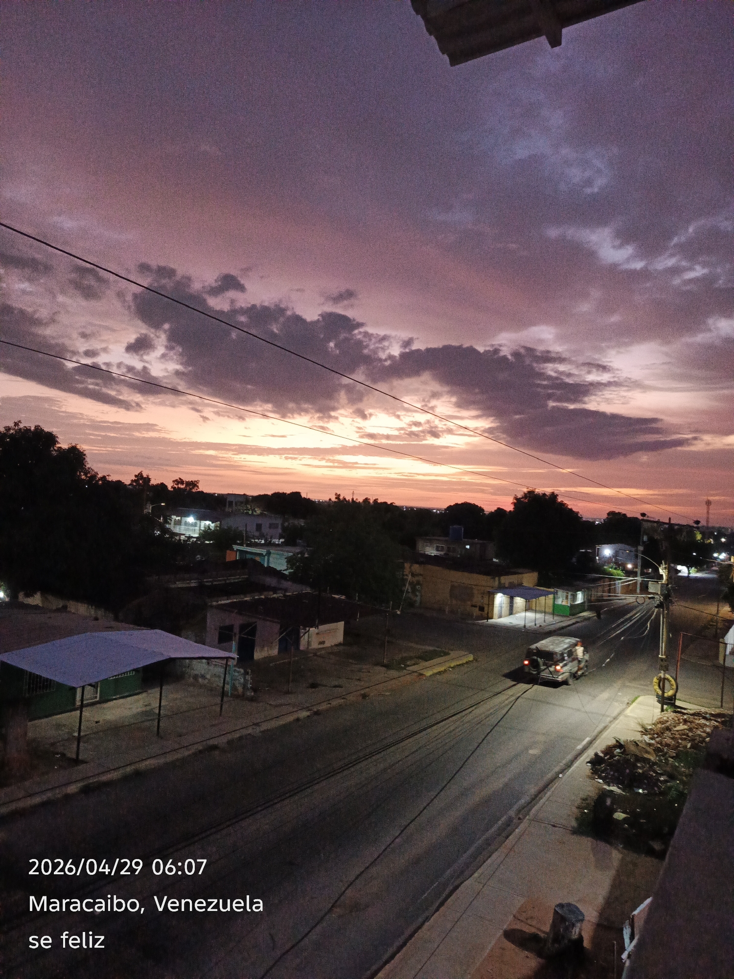 This media file presents an elevated view of an urban or suburban landscape at dawn or dusk, specifically recorded at 06:07 AM on April 29, 2026. The scene is located in Maracaibo, Venezuela, with the broader context being San Francisco, Venezuela.
The upper portion of the image is dominated by a striking sky, painted with vibrant shades of purple, pink, and orange, indicating either sunrise or sunset. Dark, scattered clouds are visible across the sky, with prominent power lines stretching horizontally across the frame.
Below the horizon, a residential area is depicted with various buildings and houses, some with illuminated windows or outdoor lights. Dark silhouettes of trees and vegetation are scattered throughout the scene. A road runs through the middle-right, where a light-colored, SUV-like vehicle is seen driving, its headlights and taillights on, casting light on the asphalt. Adjacent to the road, a pile of debris or garbage is visible. In the bottom left, there's a structure with a light-colored, angled roof, possibly an awning or a covered waiting area. A concrete or wooden stump is present in the immediate foreground. Streetlights are also visible, contributing to the artificial illumination of the scene.
Text overlays in the bottom left corner provide temporal and geographical information: "2026/04/29 06:07", "Maracaibo, Venezuela", and a positive message in Spanish: "se feliz" (be happy). The overall mood is calm and serene, capturing the quiet start or end of a day.